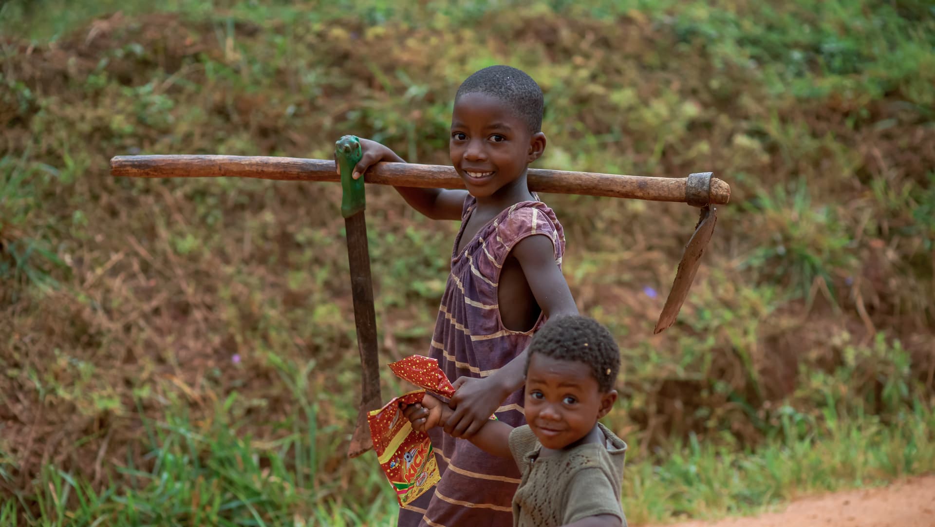 Two children walk on a path, the older one carrying a wooden yoke or stick over his shoulders.