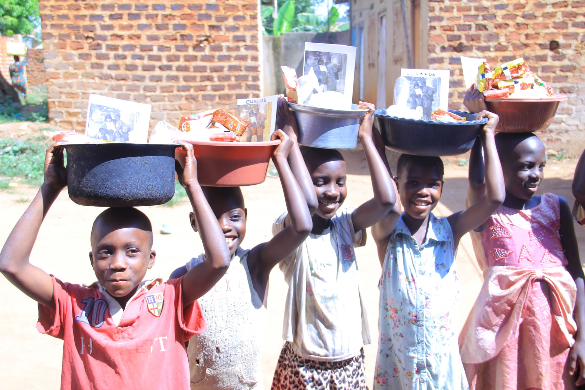 Two smiling girls walking down a dirt path carrying black basins of food on their heads.