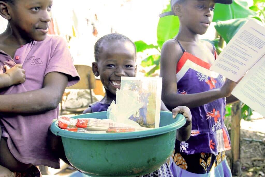 Children gather around a green basin of food items, one holding a gospel tract or paper.