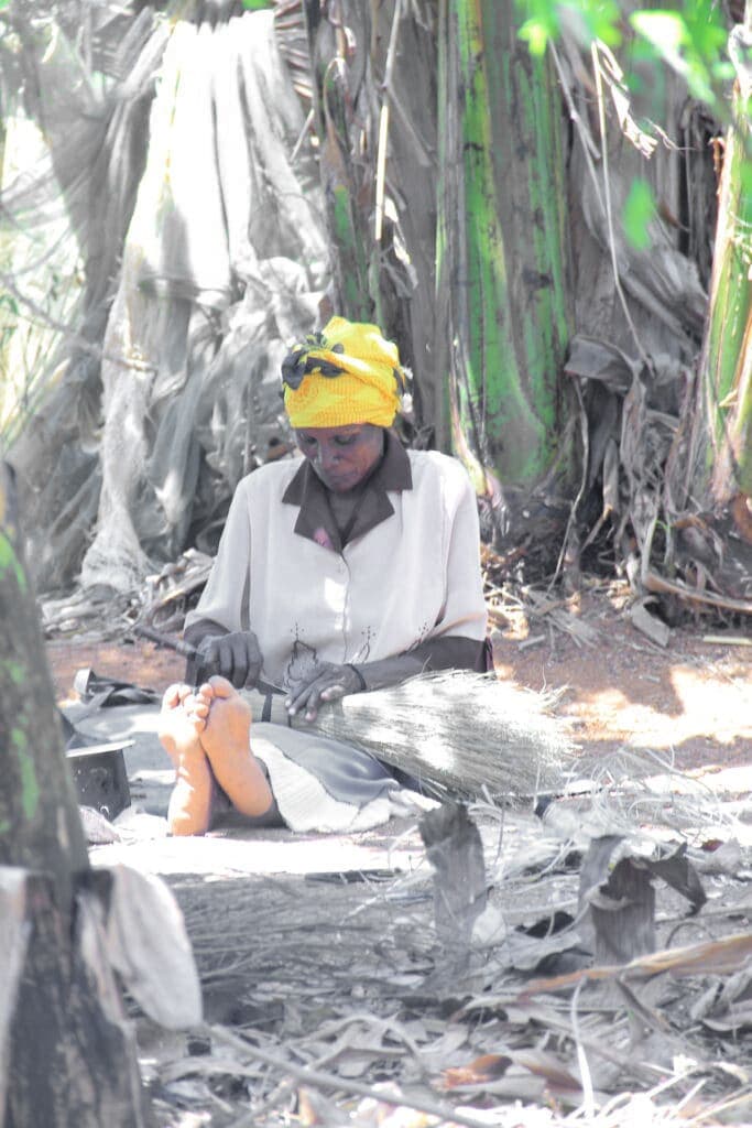 Woman in a yellow head wrap kneeling under banana leaves, working with a wooden grinding tool.