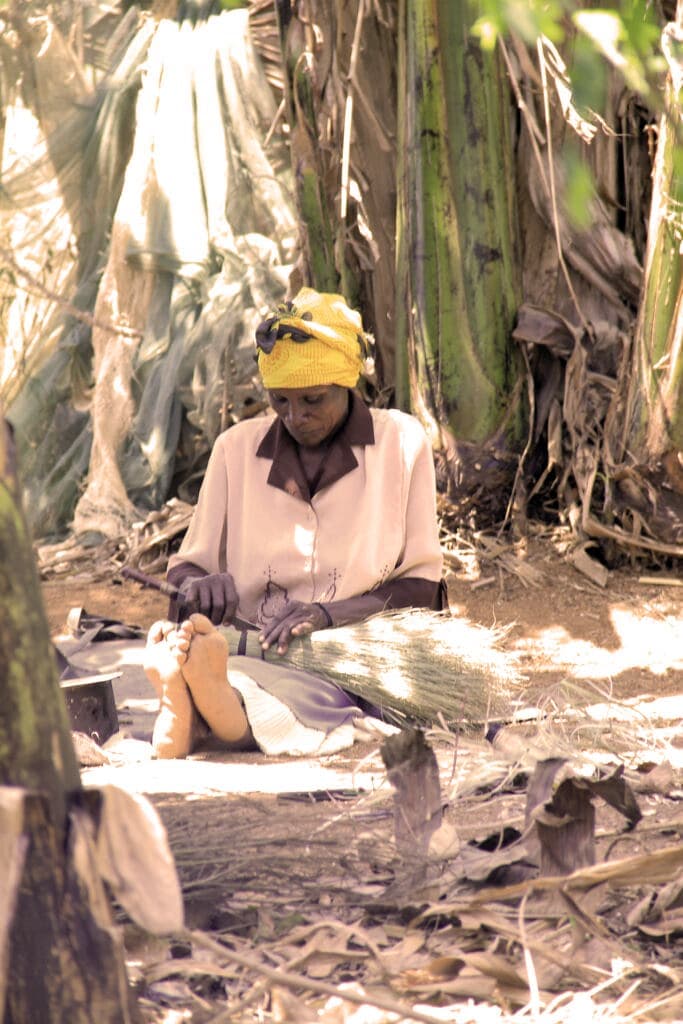 Woman wearing a yellow headscarf sits among banana plants preparing food items.