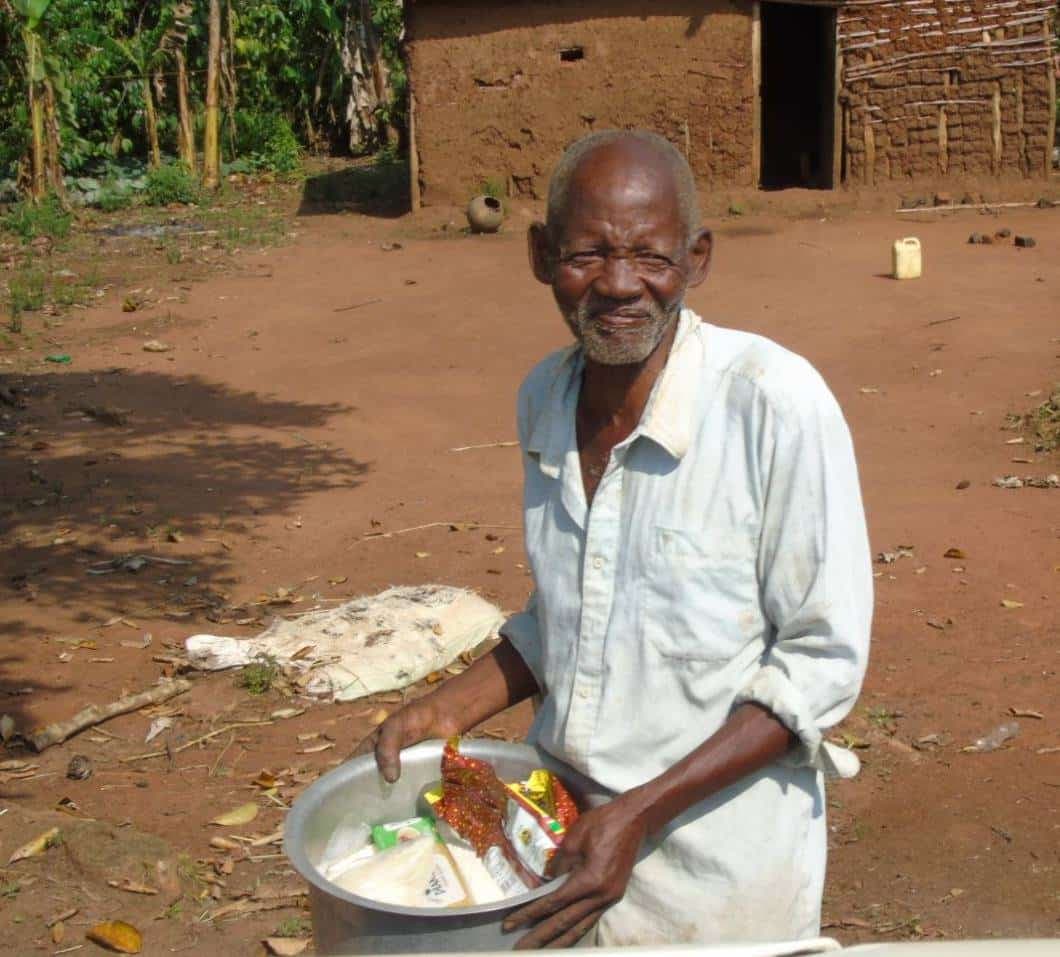 Elderly man in a white shirt standing outside a mud house holding a bowl of maize meal.