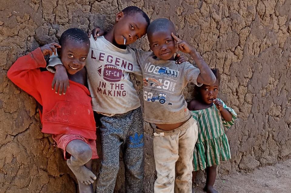 Four boys stand together against a mud wall, arms around each other, smiling.