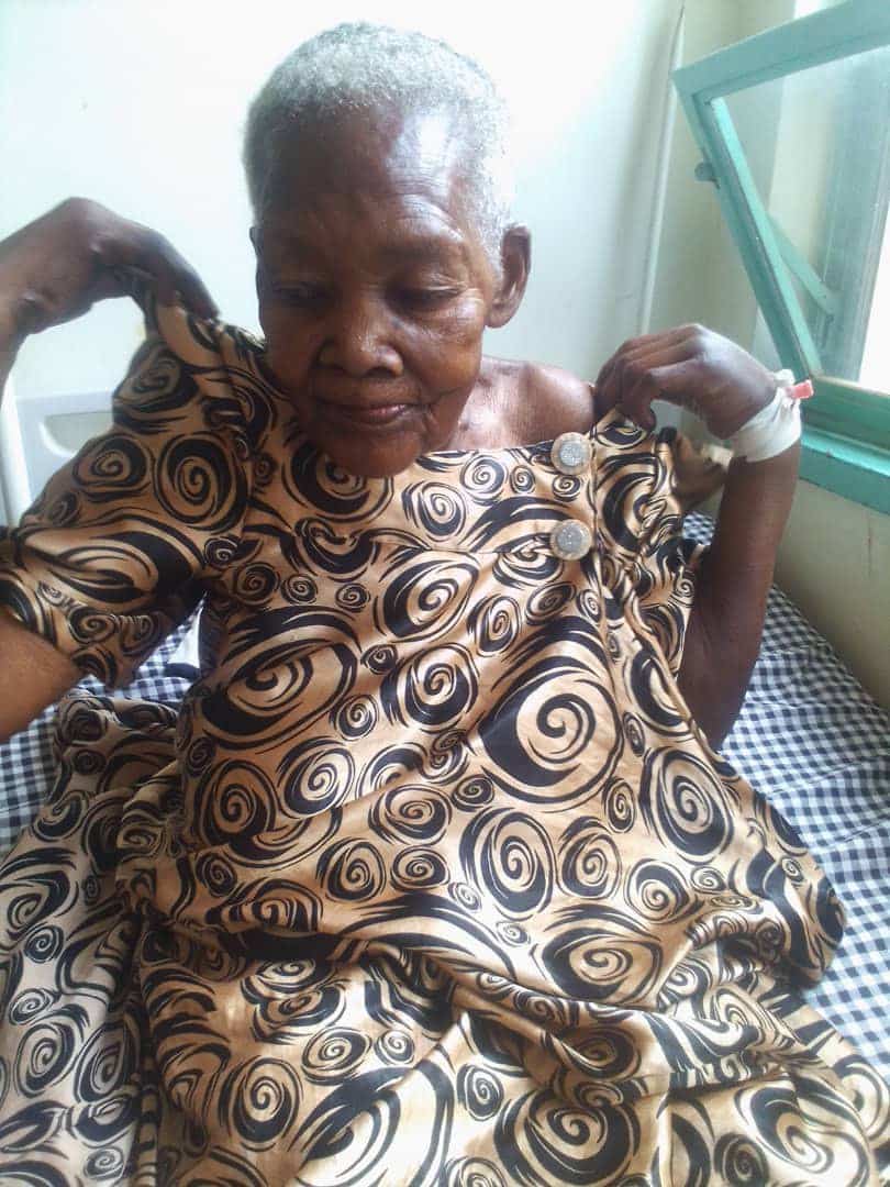 Elderly woman, Catherine Nantongo, sits on a bed adjusting her patterned dress at Miracle Medical Center.