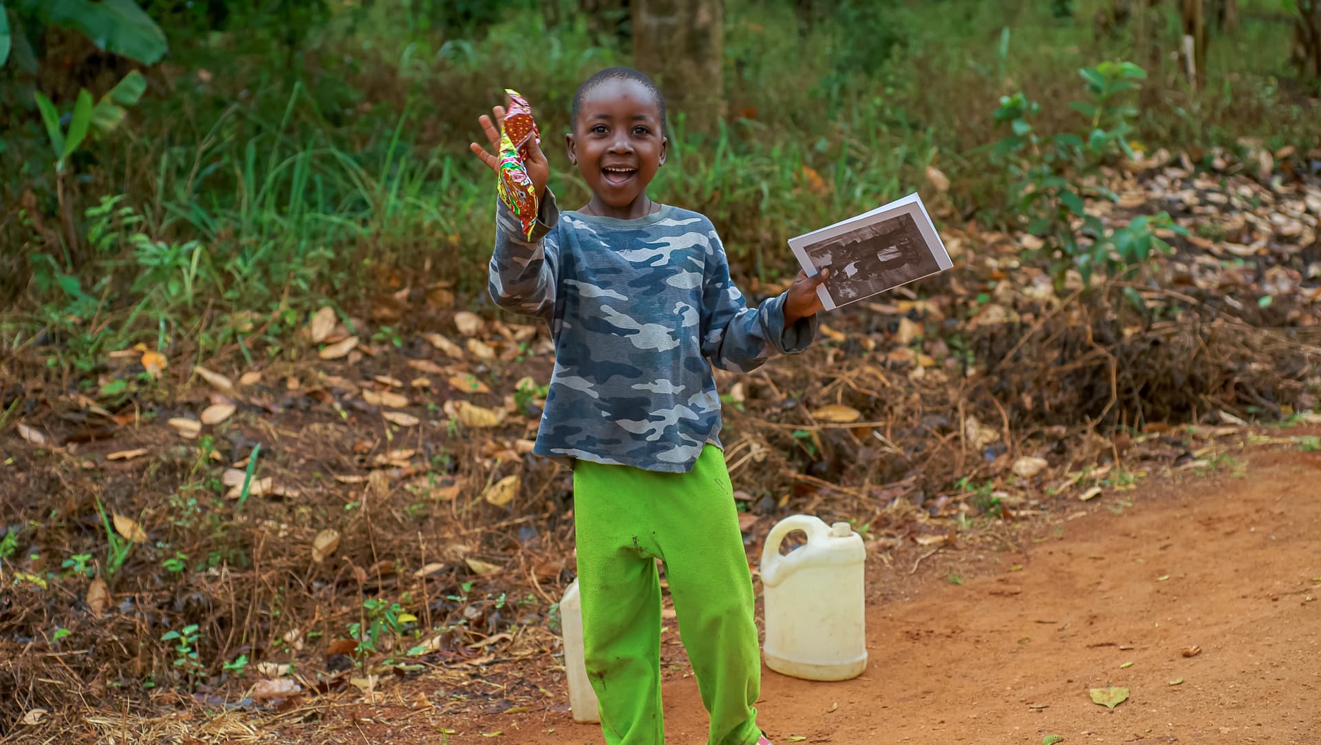 Young boy in green pants smiles and gestures while holding a gospel tract, a water jerrycan beside him.
