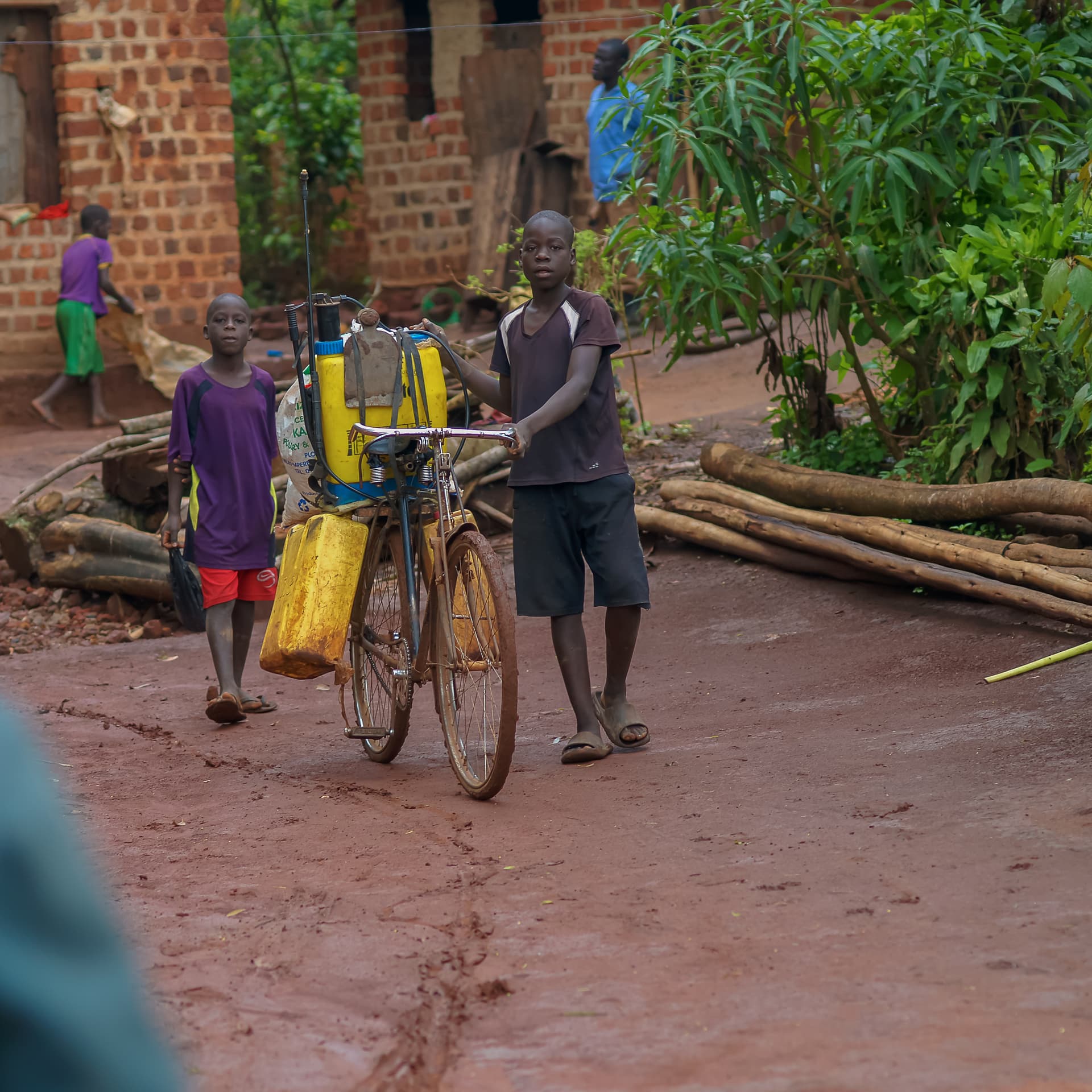 Two boys pushing a bicycle loaded with yellow jerrycans of water along a dirt path near a brick building.