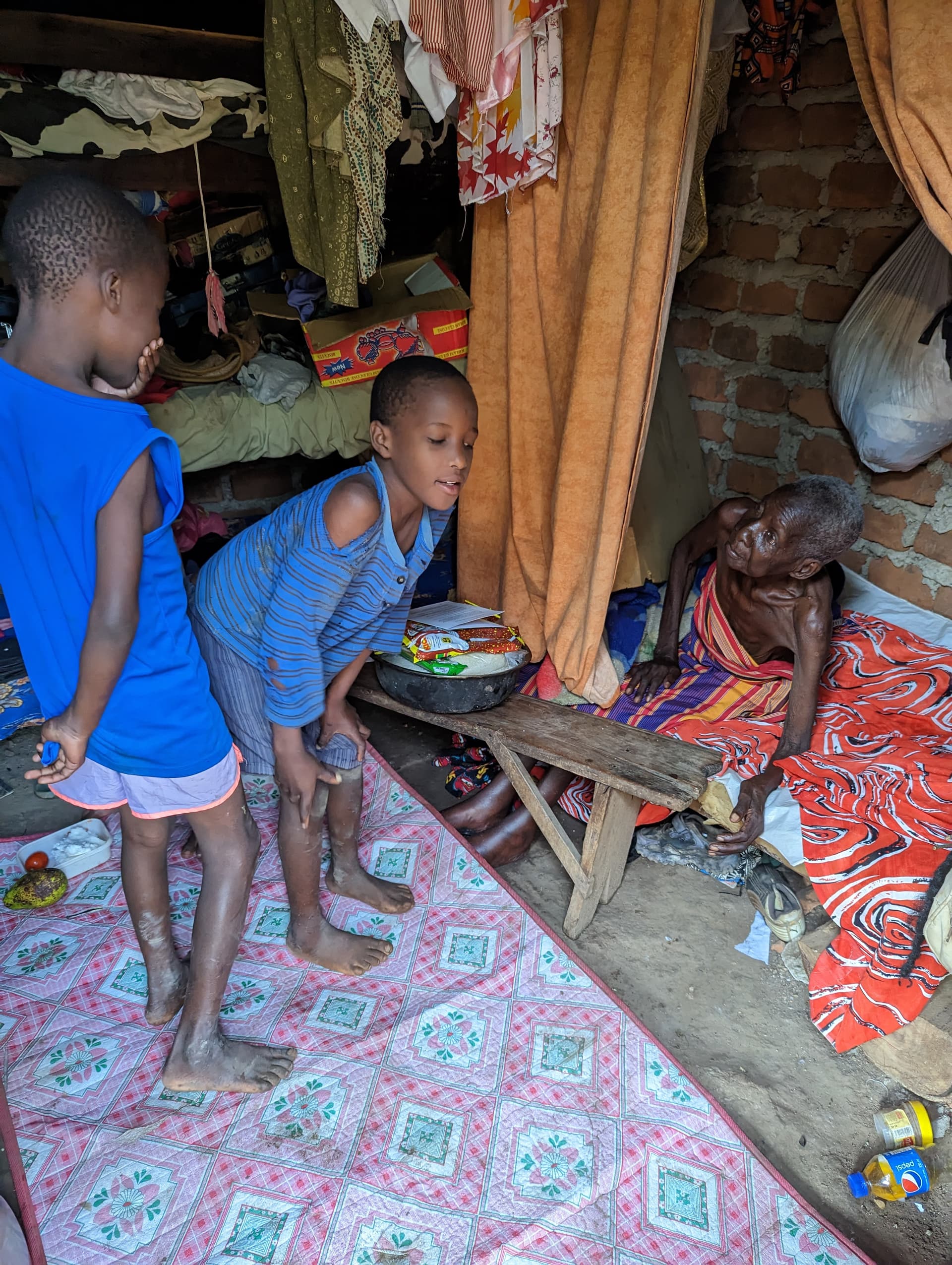 Two children standing beside an elderly woman resting on a bed inside a home with colorful curtains.