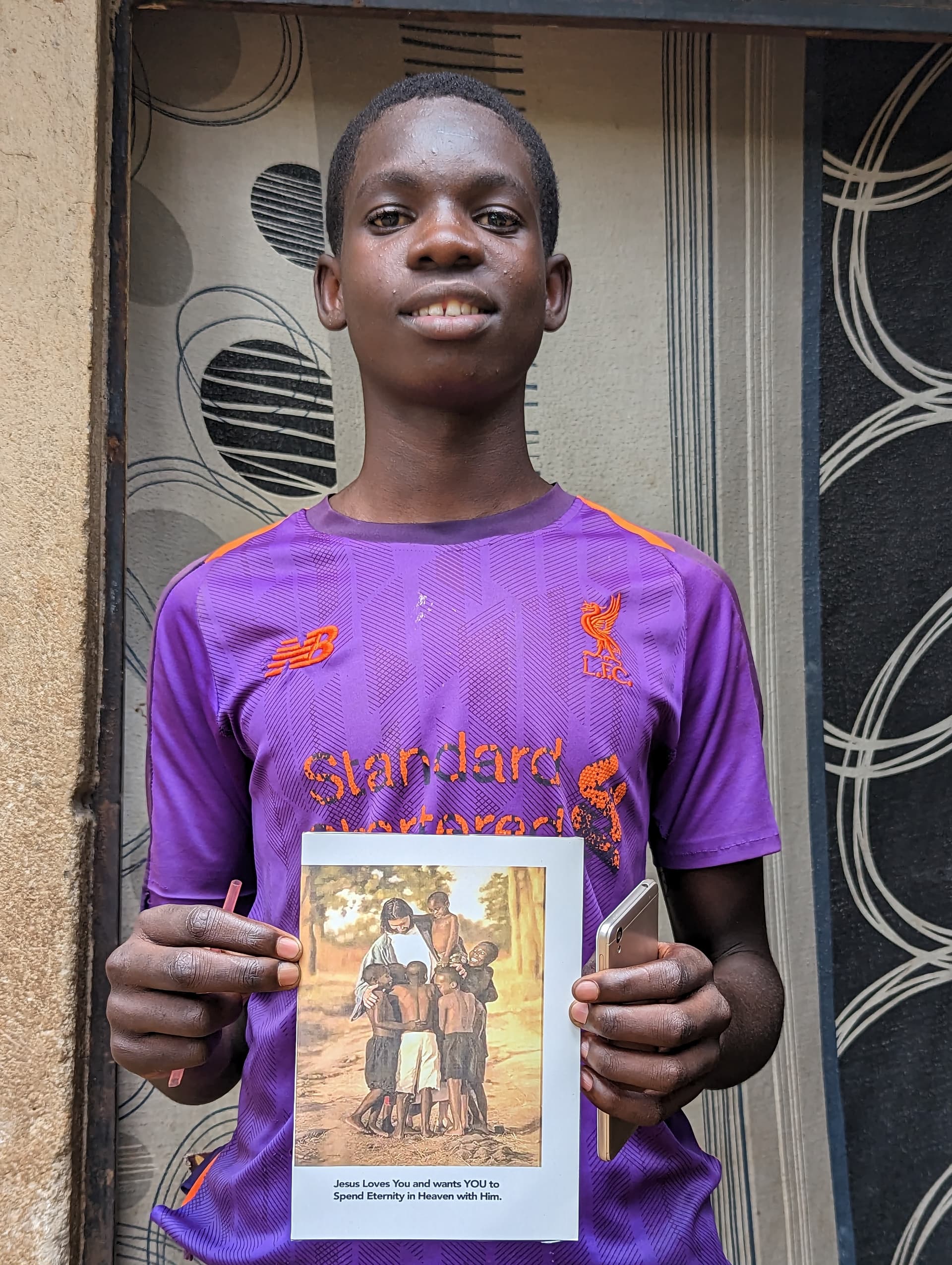 Teen boy in a purple school shirt holding up an illustrated storybook at a doorway, JFM-sponsored student Joseph.