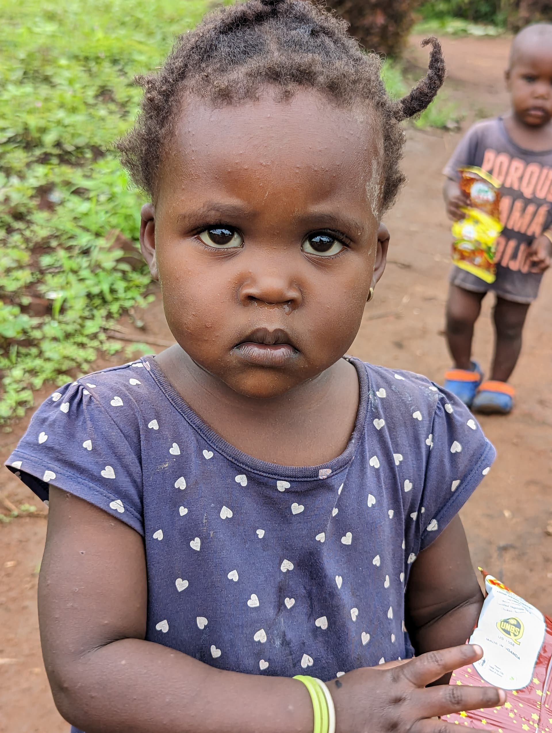 Toddler girl in a polka-dot dress holding a small item, with other children carrying food gift bags behind her.