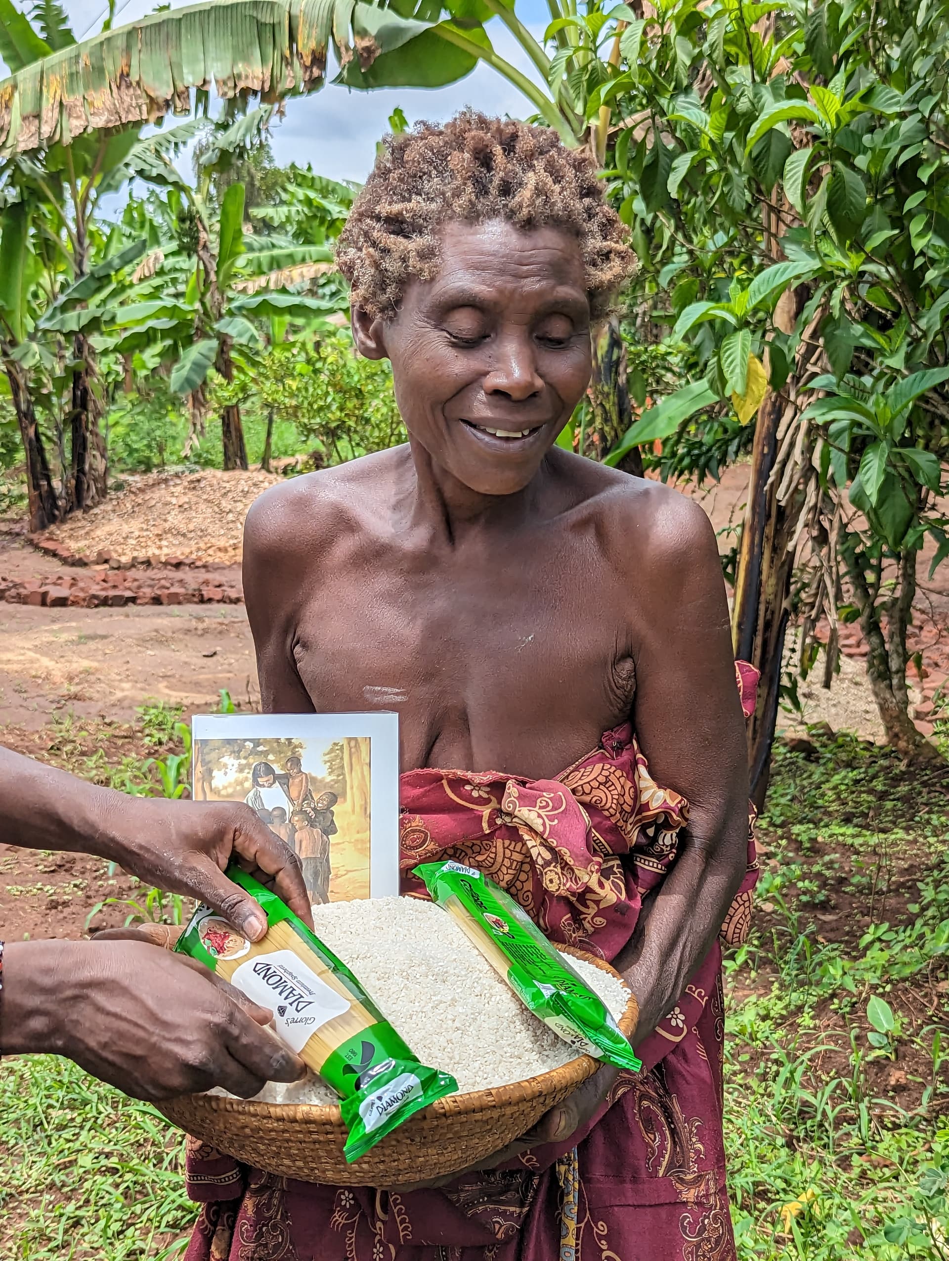 A thin elderly woman stands in a banana grove holding a food packet and photo tract.