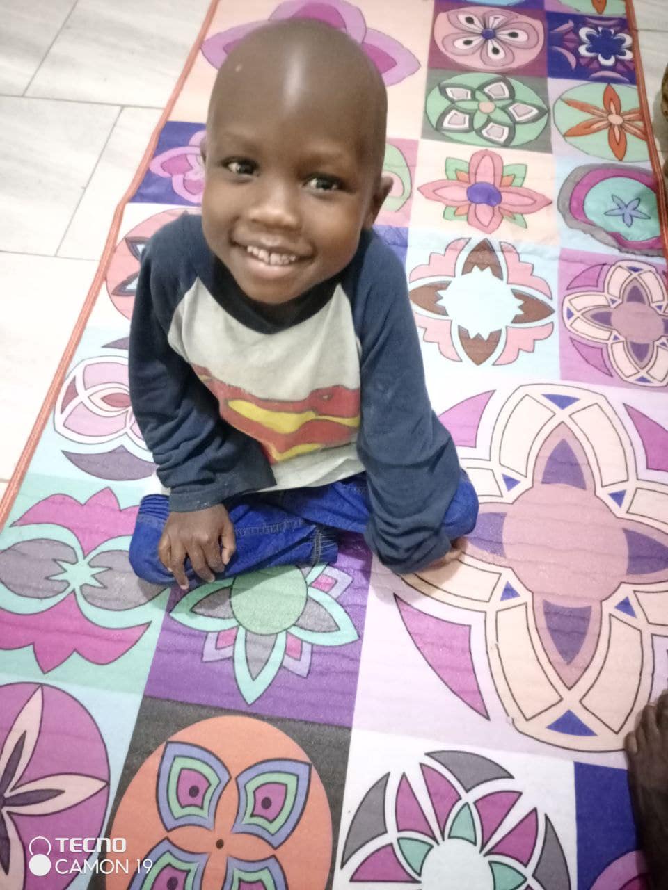 Young boy sits smiling on a colorful patterned mat indoors.