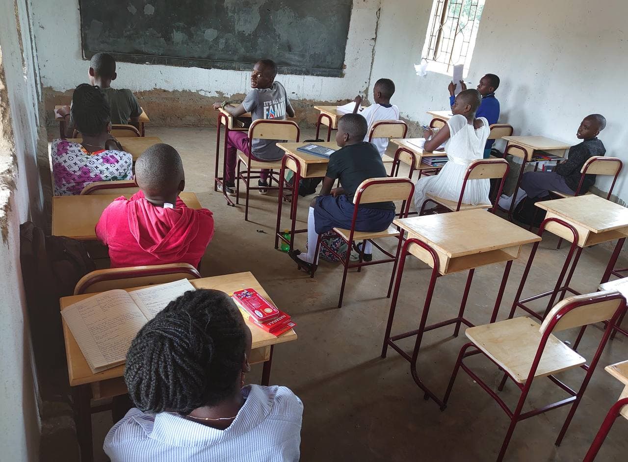 Students sit at newly delivered classroom desks and benches beneath a chalkboard at Greater High School.