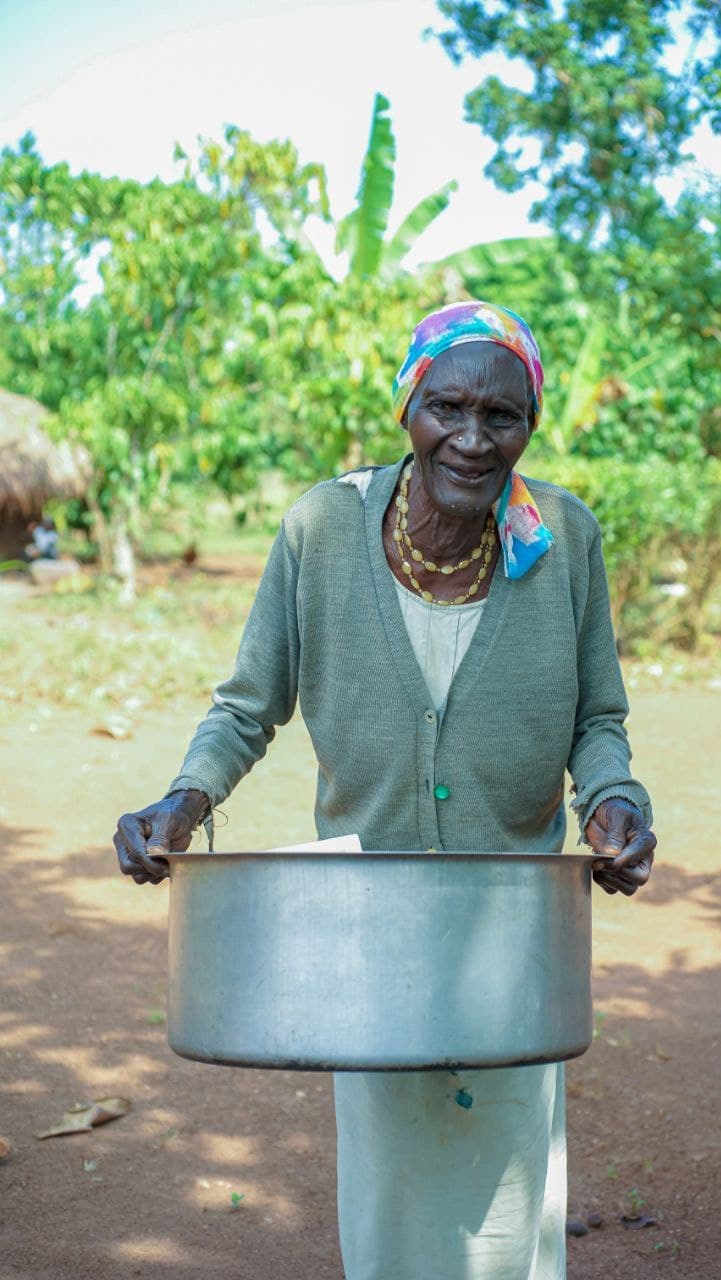 Elderly Ugandan woman in a green cardigan holds an empty metal basin outdoors near banana leaves.