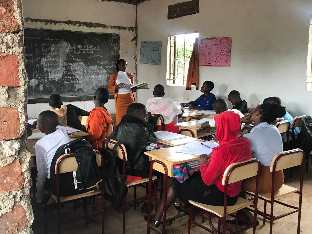 Teacher standing at a chalkboard in front of students seated at desks in a Ugandan classroom.