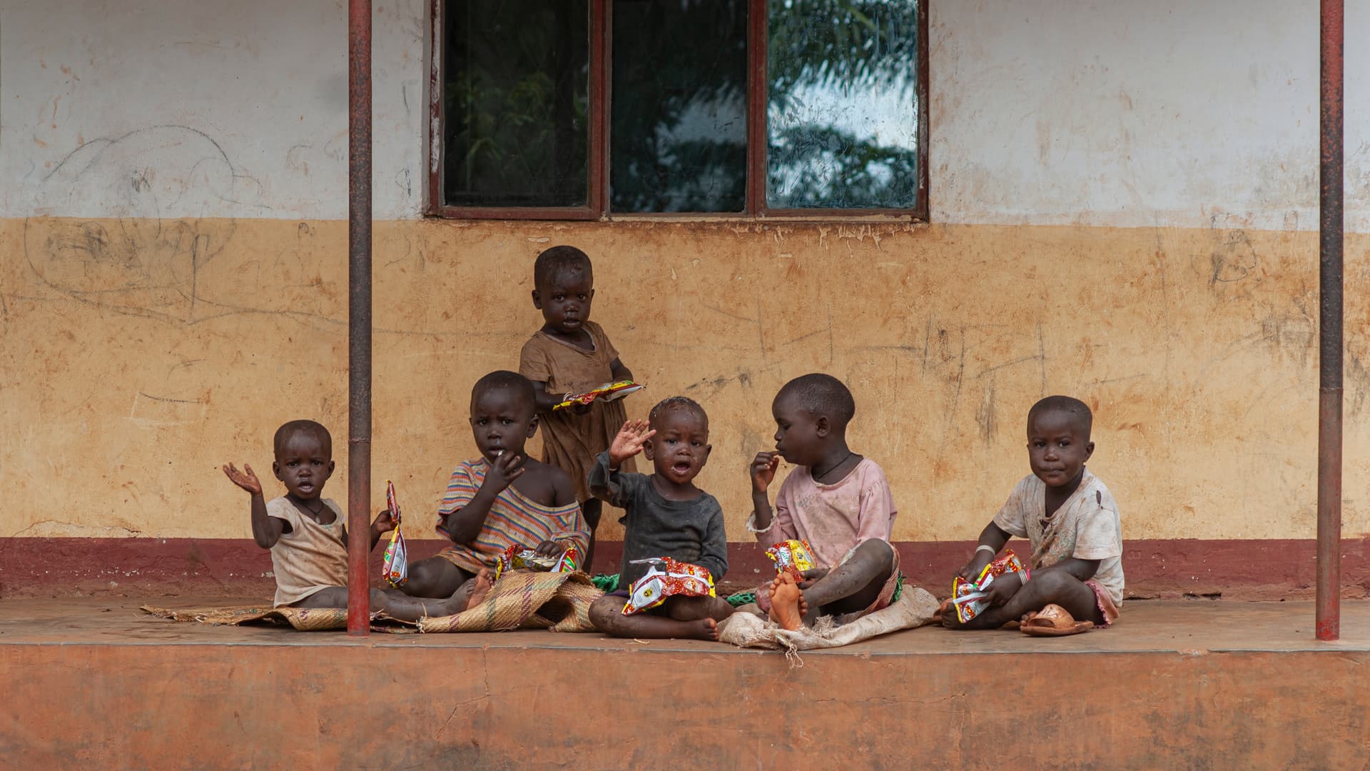 Several children sit together on a porch bench outside a building.
