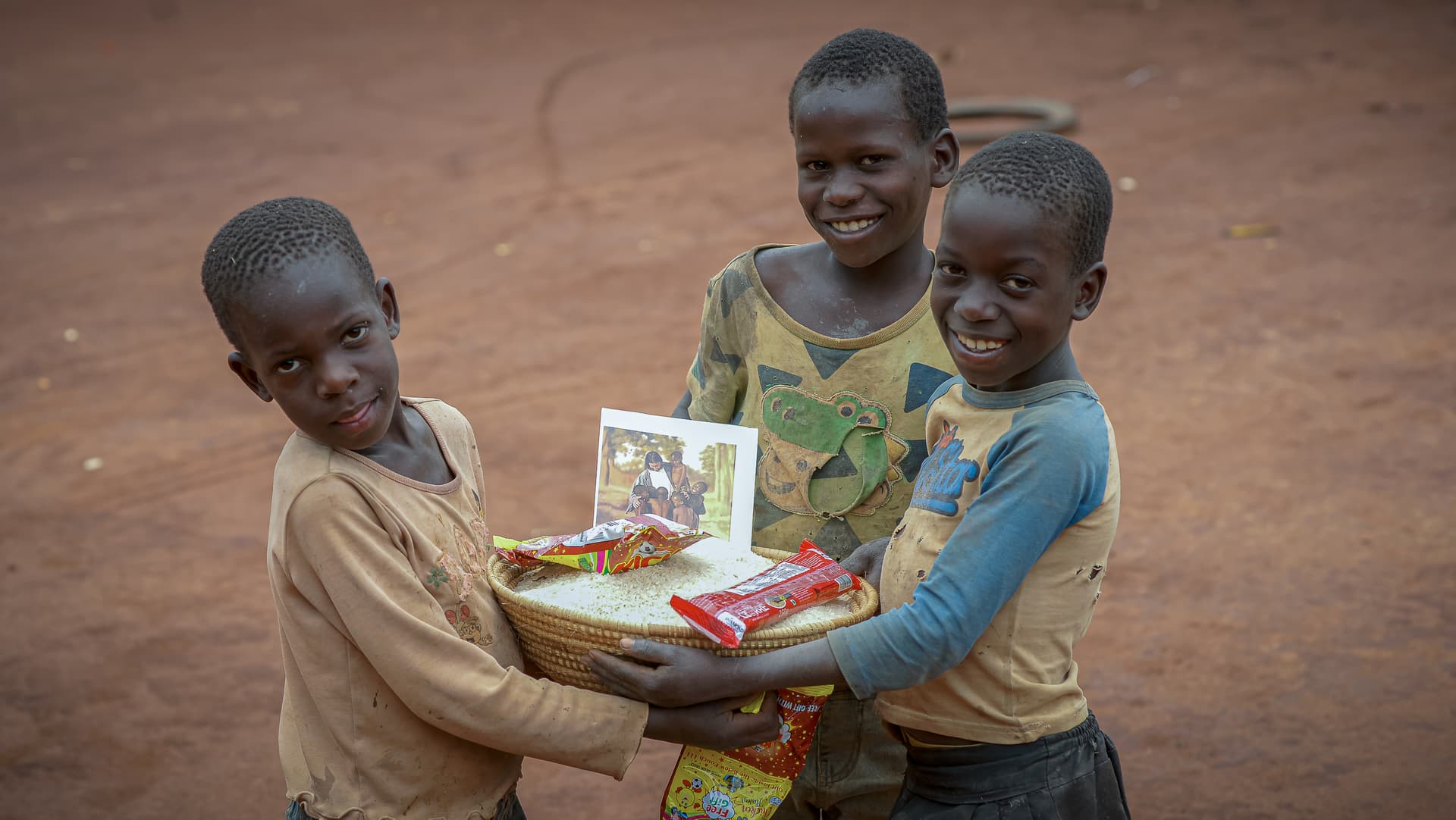 Three children stand together with arms linked, holding a food package with a photo tract.