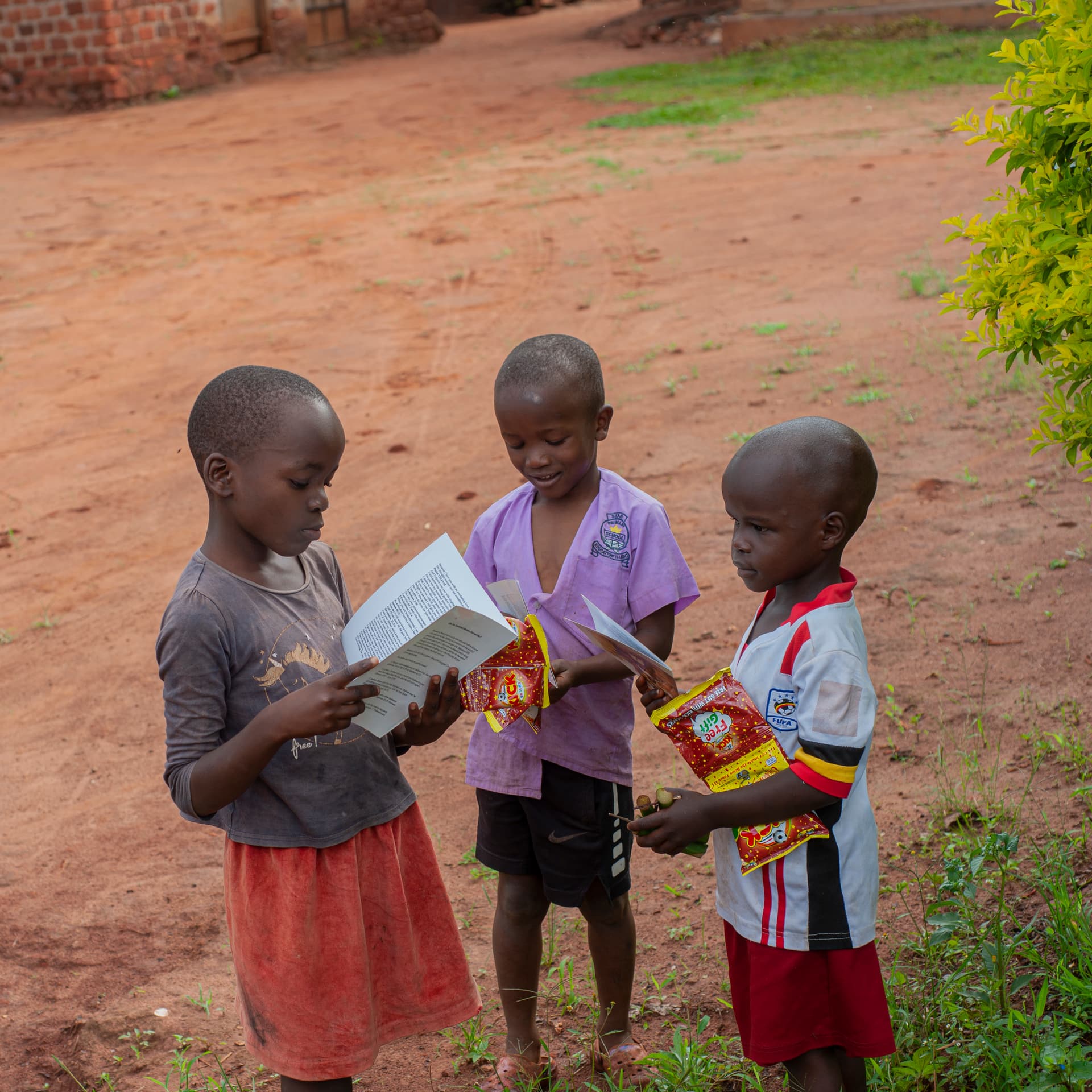 Three children stand together on a dirt path reading a book or tract, two boys and a girl in a floral dress.