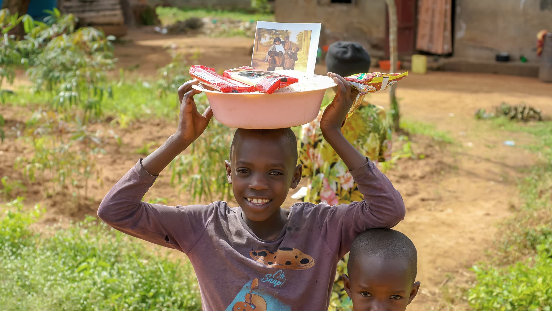 A girl balances a red basin holding food and a photo tract on her head, standing beside a younger child.