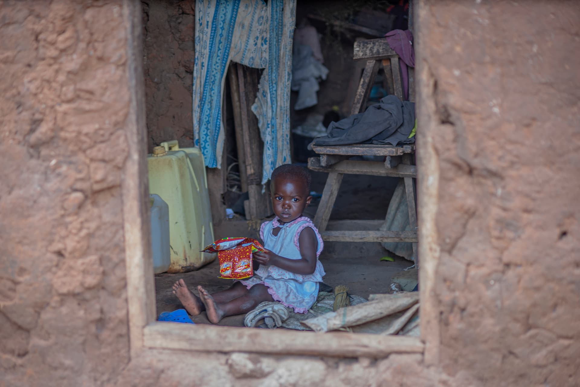A young child sits inside the window opening of a mud-brick home in Uganda, laundry draped nearby.