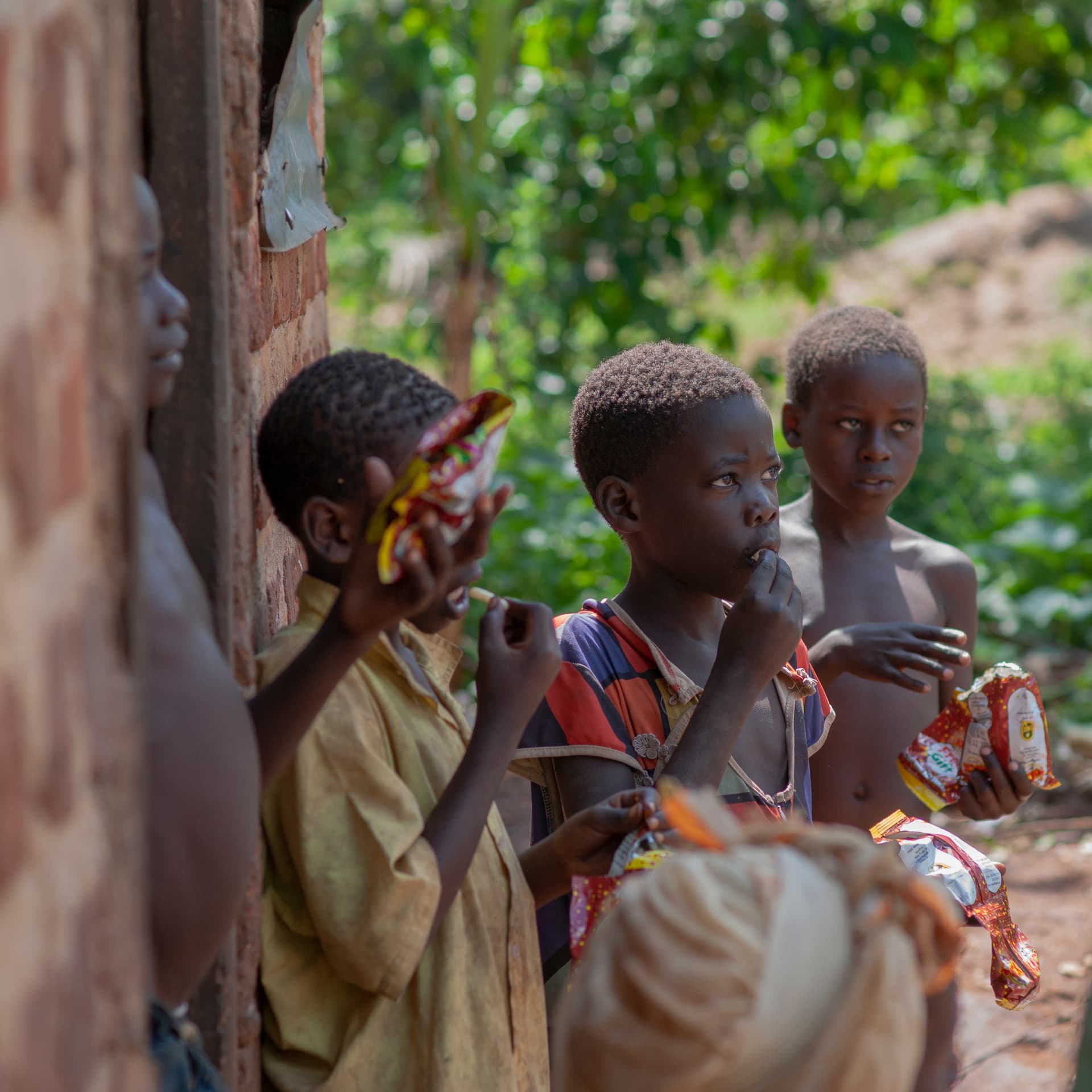 Children stand near a wooden structure outdoors, one holding a bottled drink.