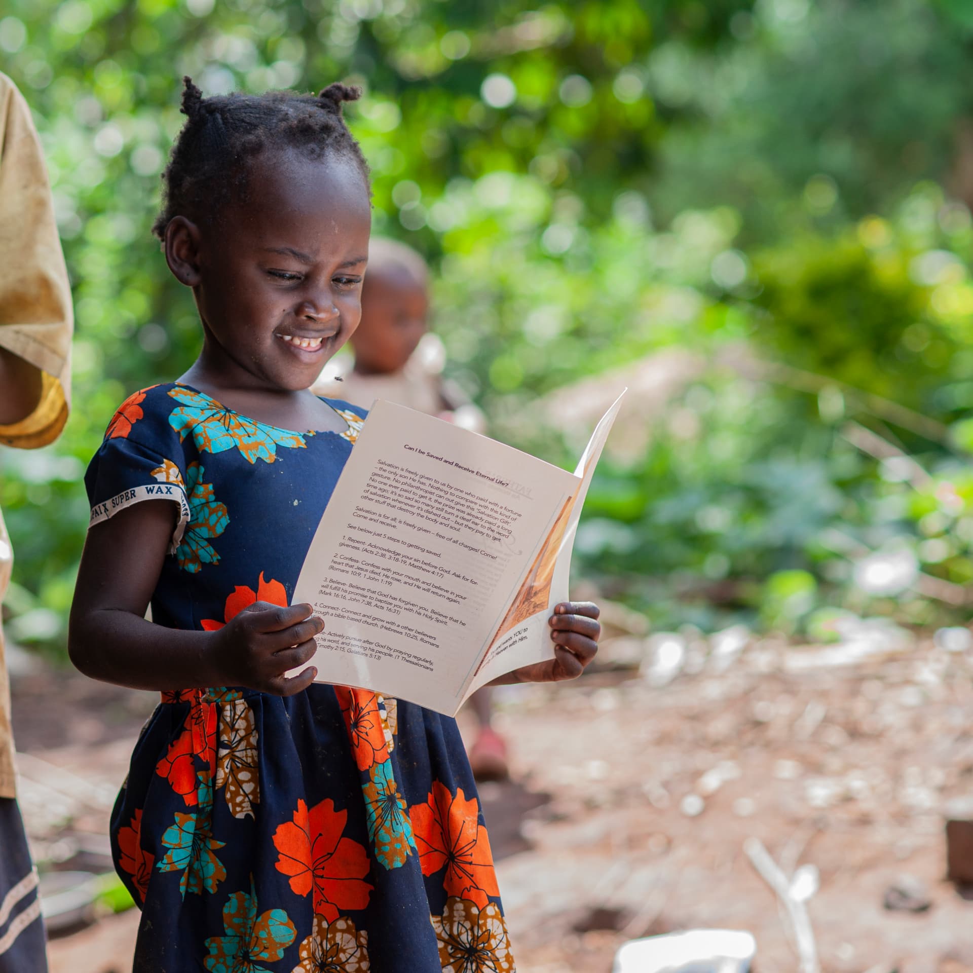 A girl in a floral dress smiles while reading a gospel tract outdoors.
