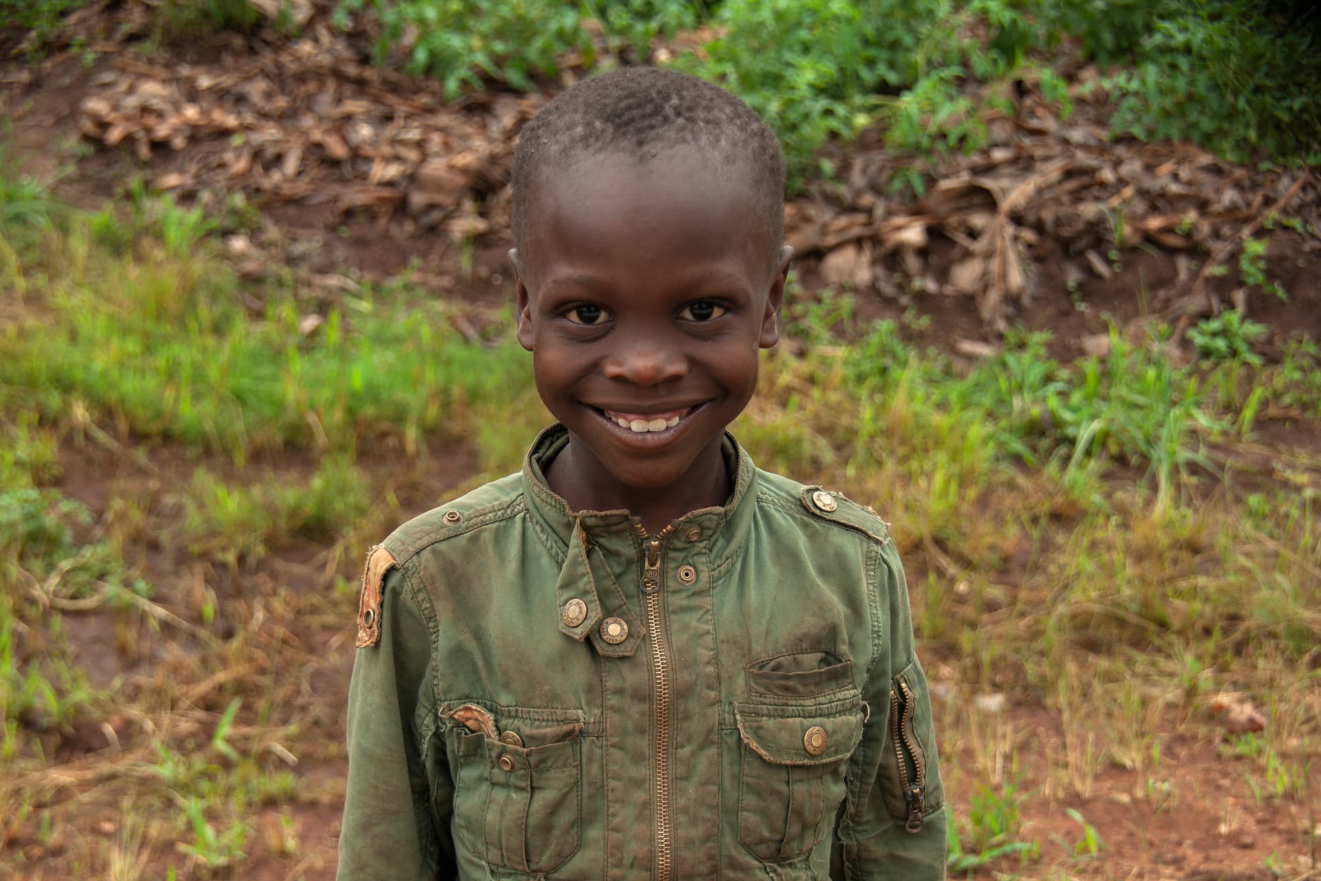 A smiling boy in a green jacket stands outdoors near a hillside.
