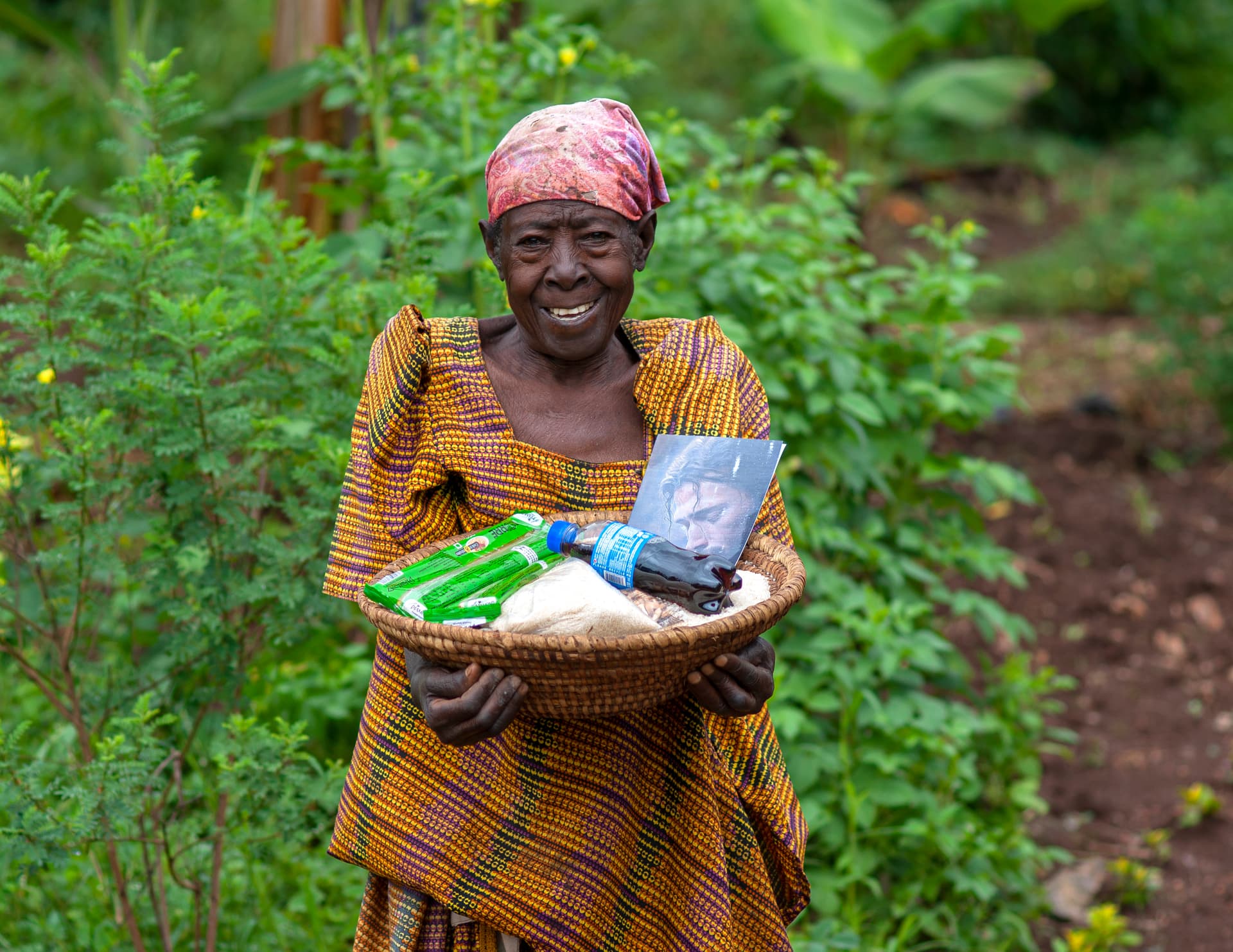A woman in a headscarf smiles while holding a basket of food and a photo card in a green field.