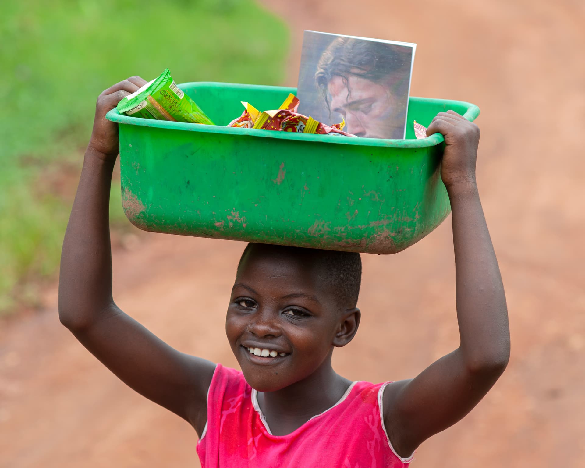 A girl in a red top carries a green bowl overhead with a photo card inside.