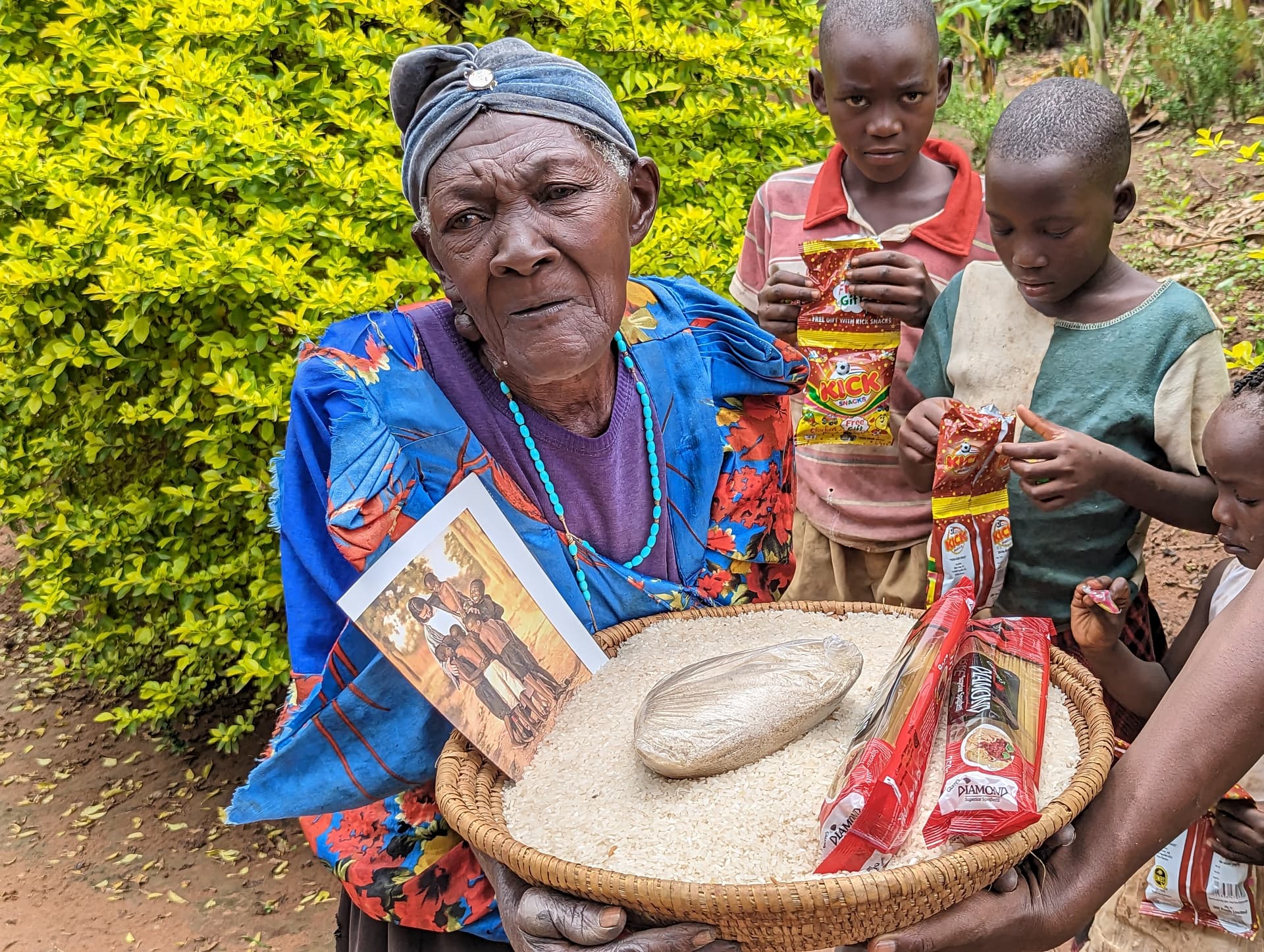 An elderly woman surrounded by several children holds a basket of food packets and a photo tract outdoors.