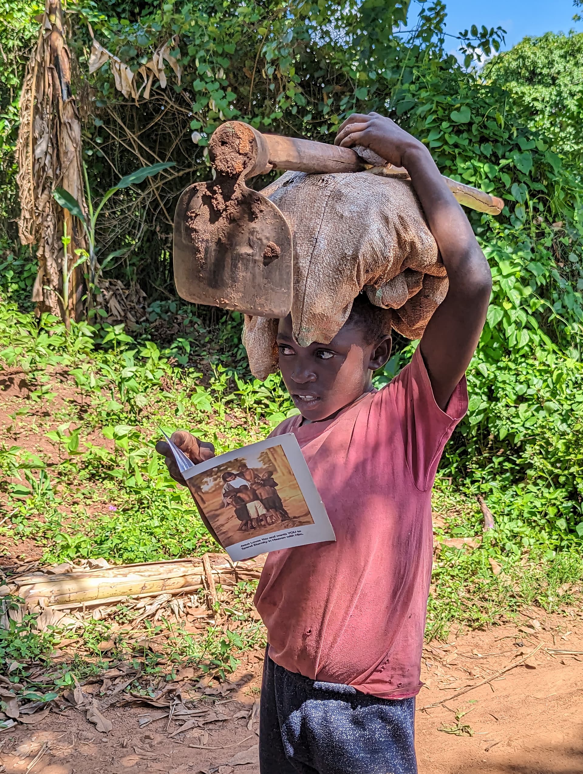 A boy carries a large bundle on his head while holding open a tract book, walking along a green path.