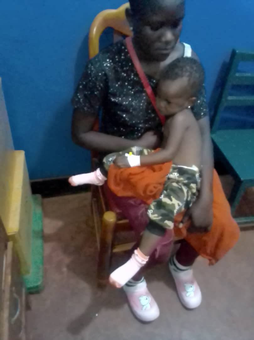 A man holds a young child wearing an orthopedic leg brace and white shoes, seated in a blue-walled clinic room.