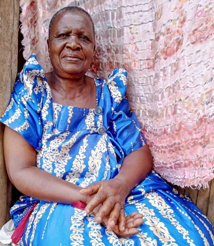 An elderly woman in a blue and white patterned dress sits indoors near a curtain.