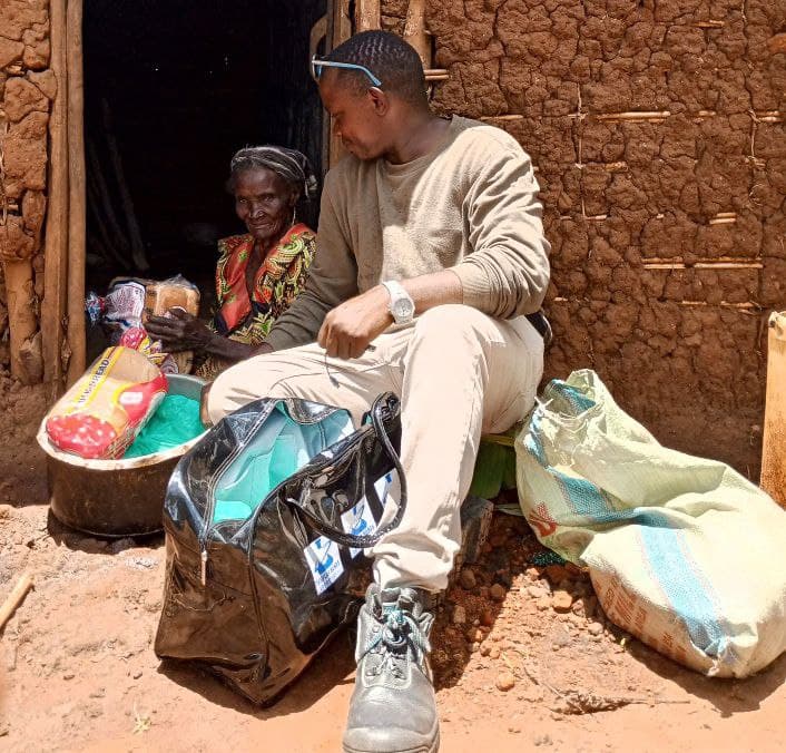 An elderly woman sits while a young man hands her sacks of food outside a mud-brick home.