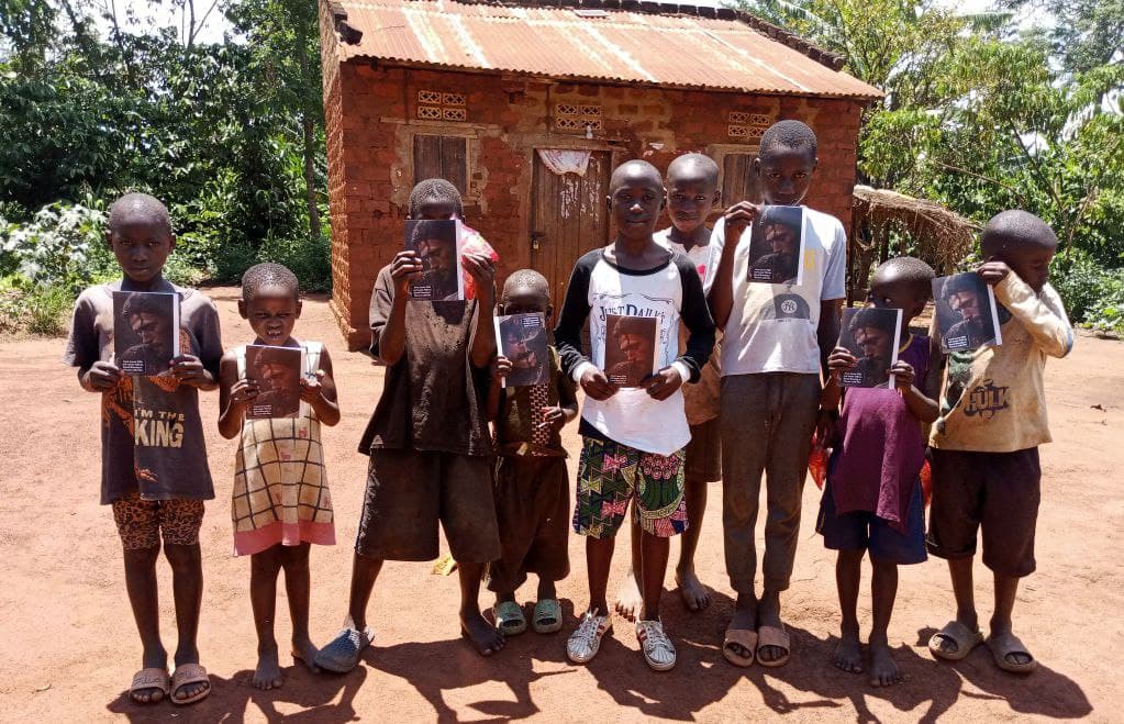 A group of children stand together outside a brick house holding gospel tracts and food items.