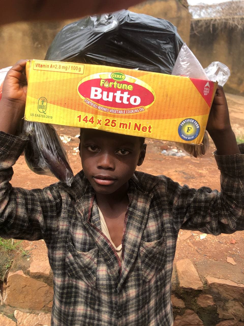 A boy holds up a large yellow package of cooking oil, standing outdoors on bare dirt ground.