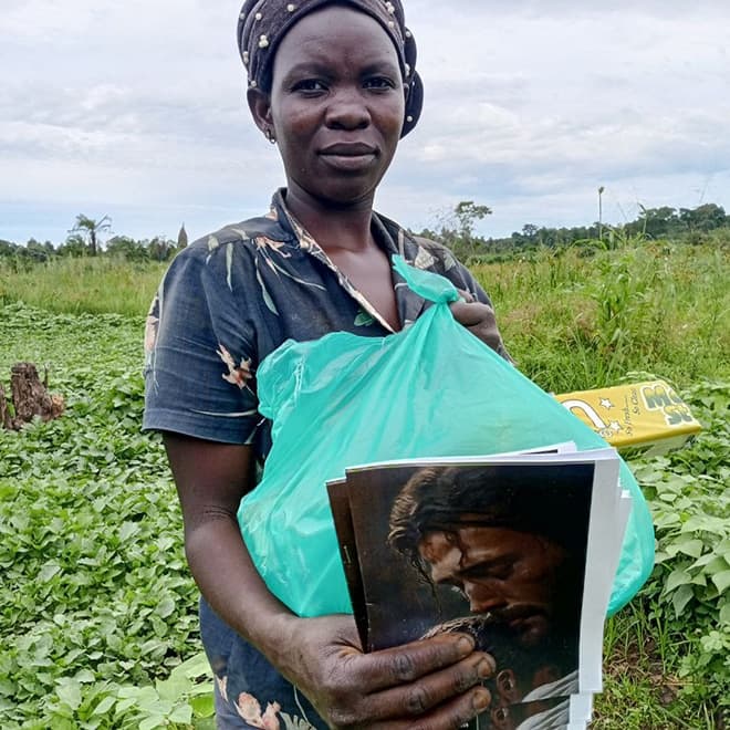 Nassazi Kyolaba stands in a farm field holding a green bag with photo cards inside.