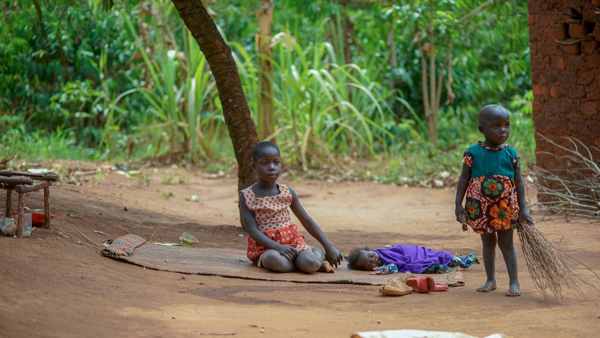 Two young children sit together under a tree next to a mat with belongings.