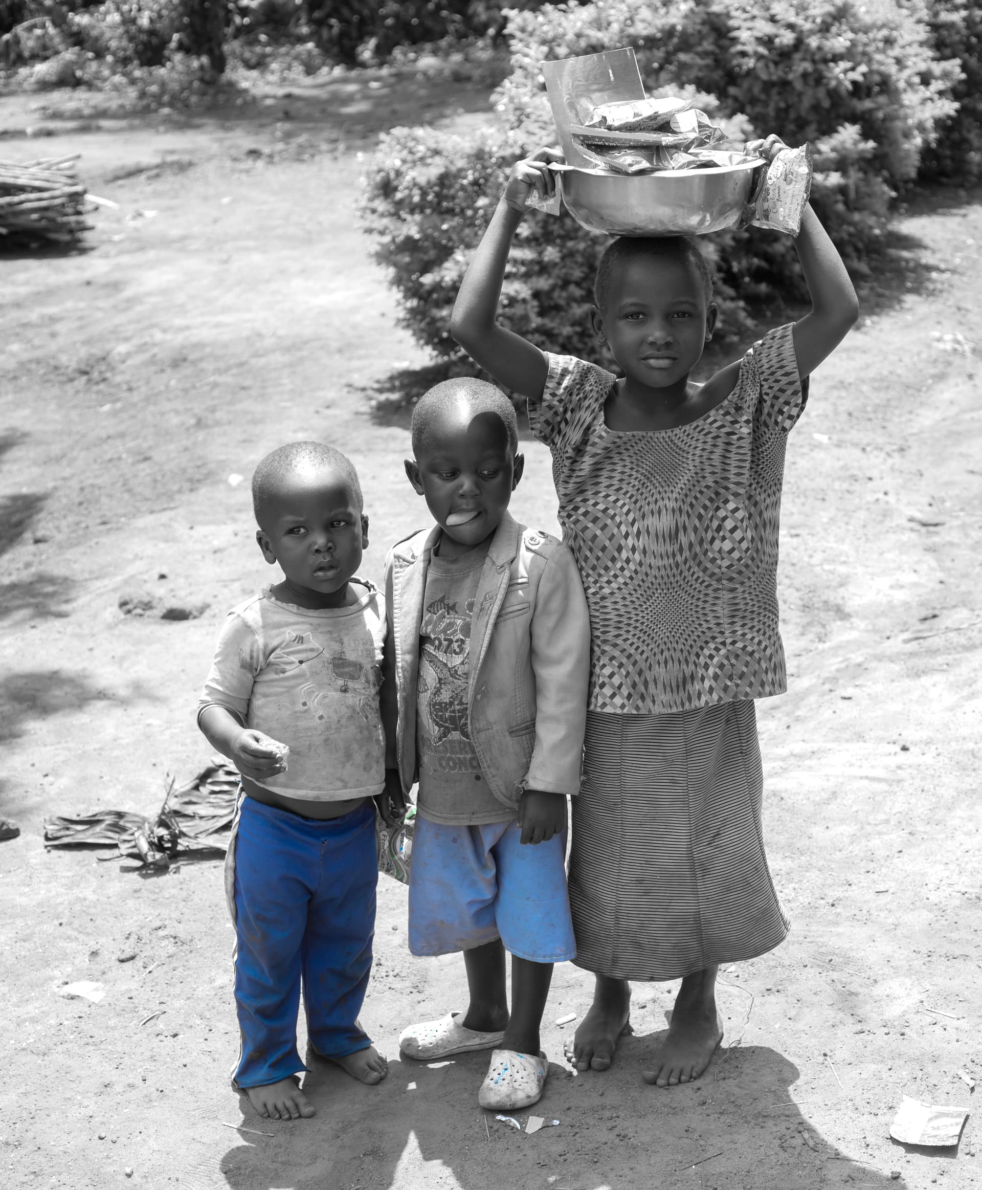 Black-and-white photo of a girl carrying a basin on her head with two younger boys standing beside her.