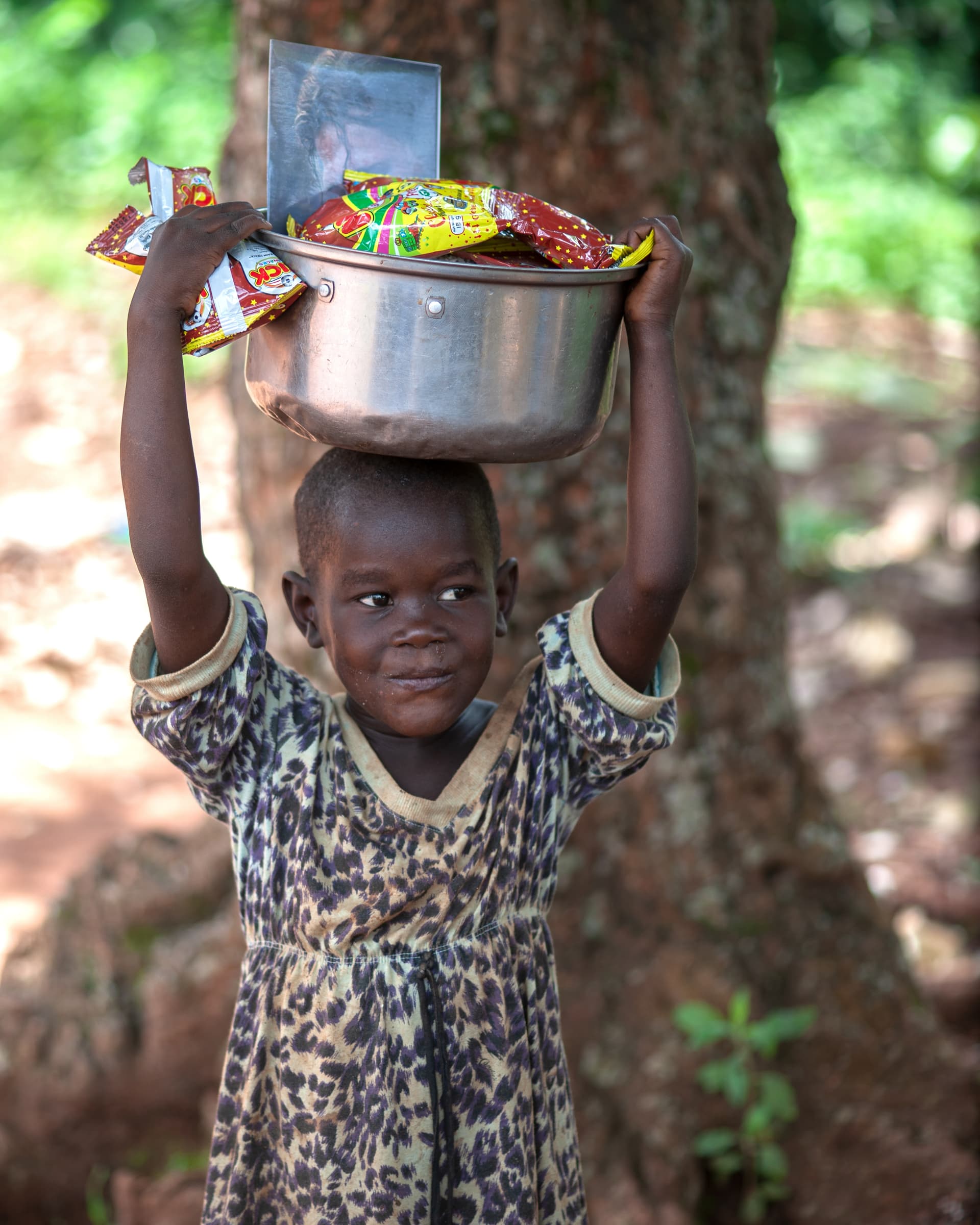 A boy in a leopard-print shirt lifts a silver pot of food onto his head.