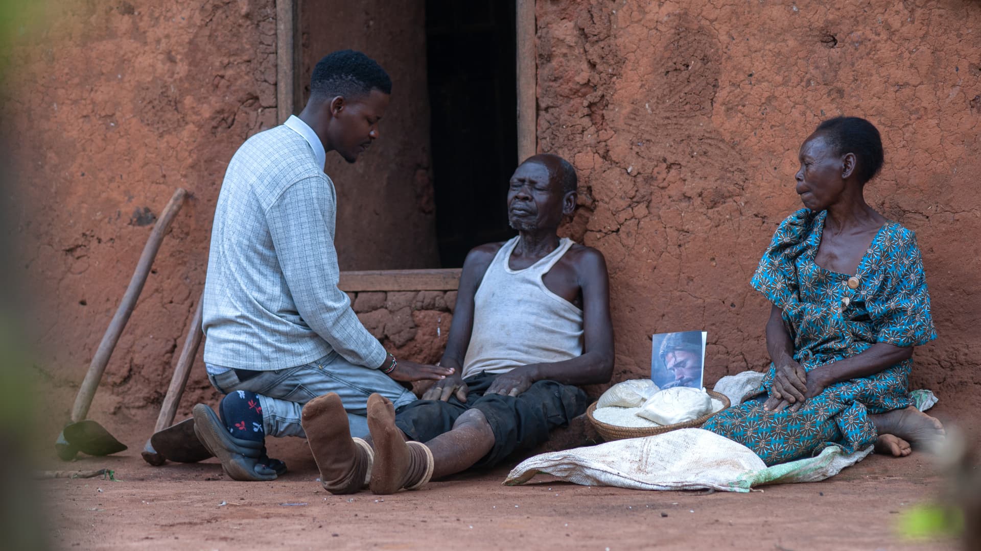 A JFM volunteer kneels to talk with Ssali Salpha and a woman seated outside a mud-brick home.