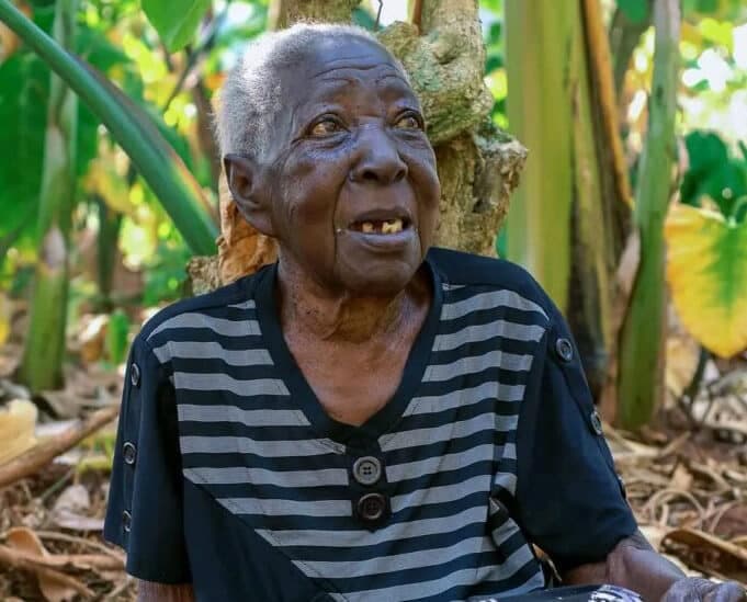 An elderly woman stands among banana plant leaves wearing a striped shirt.
