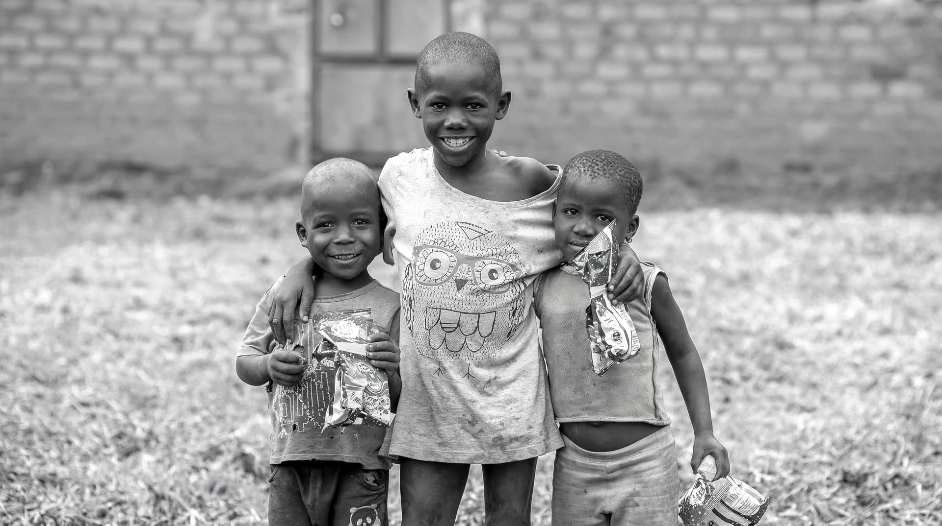 Black-and-white photo of a boy with his arm around two younger children, holding small items, outside a brick building.