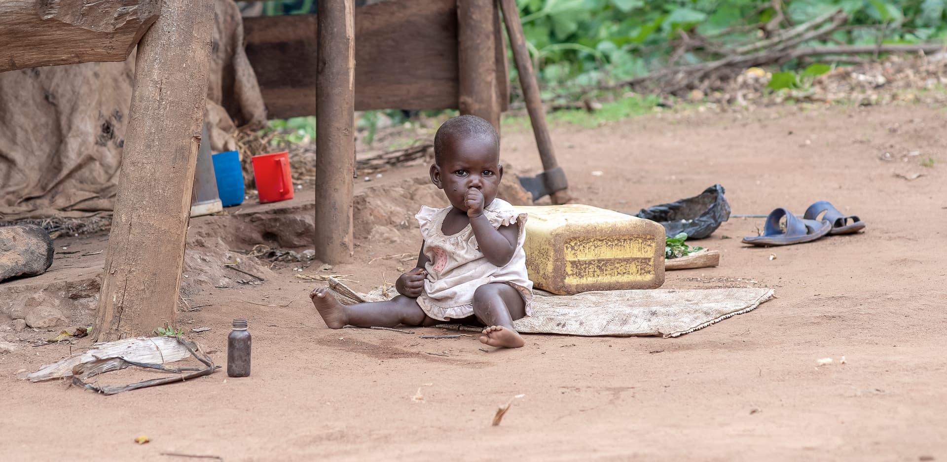 Small children sit on the ground outside a wooden structure with food items and sandals nearby.