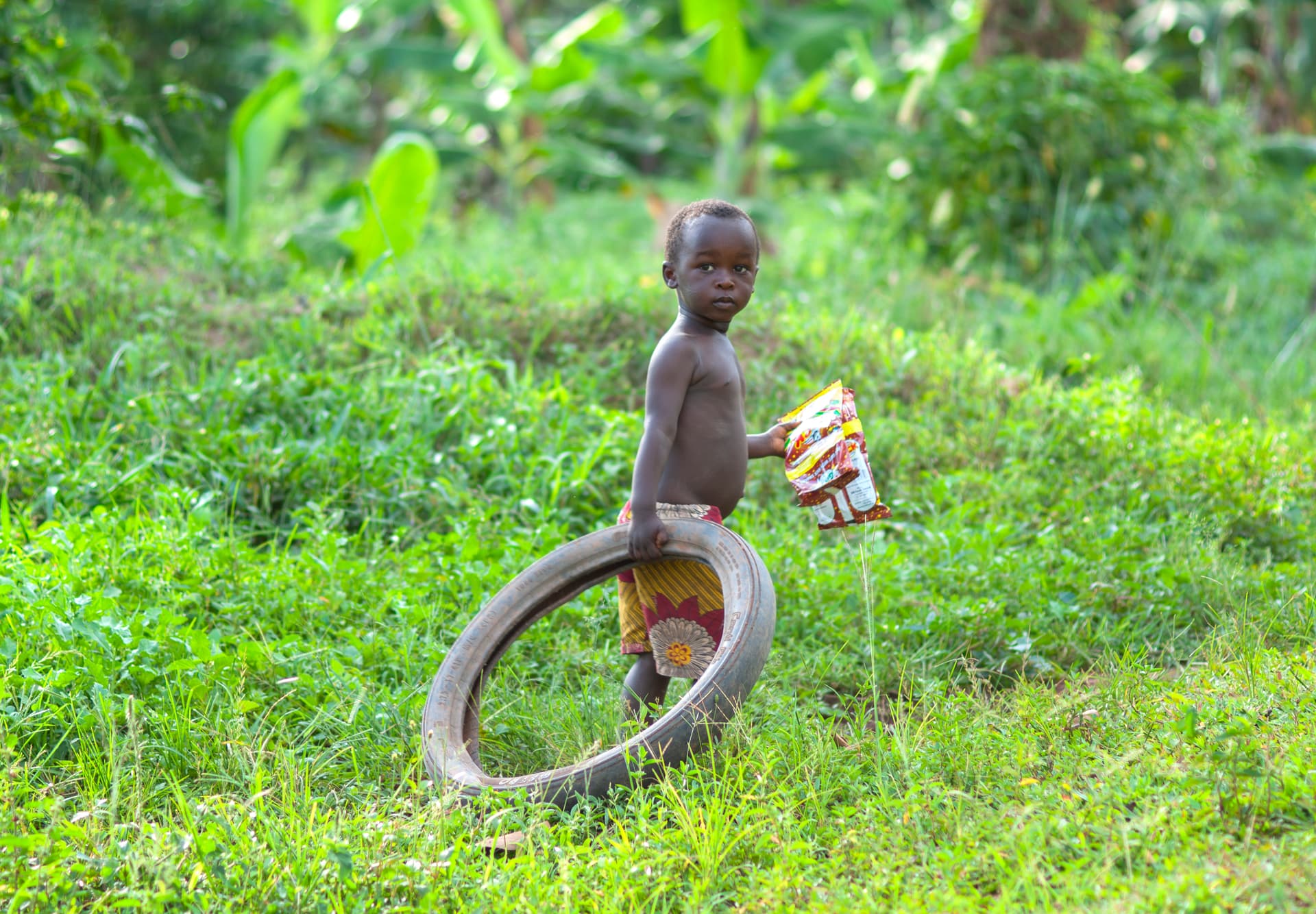 Child standing in tall grass holding an old tire and a food packet