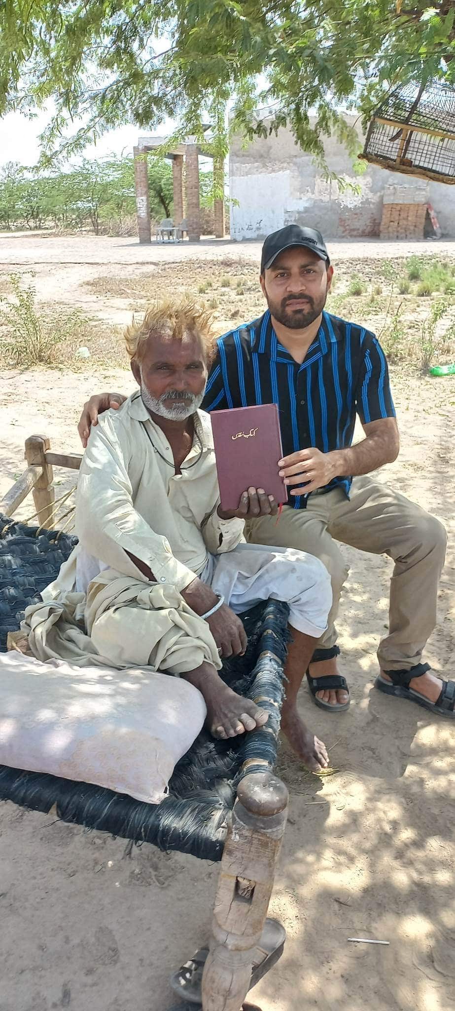 An older man seated on a bench and a younger man stand together holding a Bible outdoors.