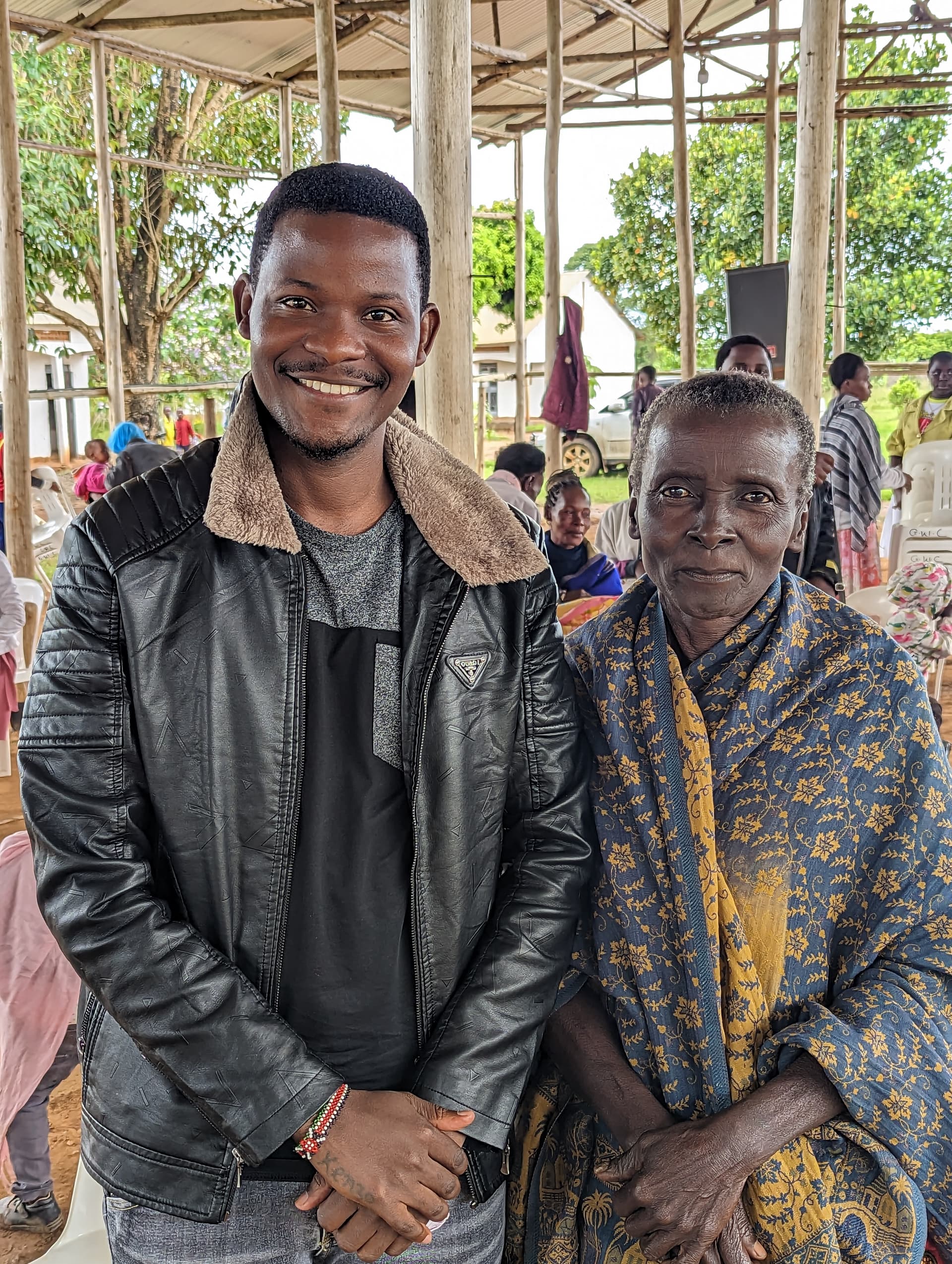 A man in a leather jacket stands arm in arm with an elderly woman in a patterned outfit under a shelter.