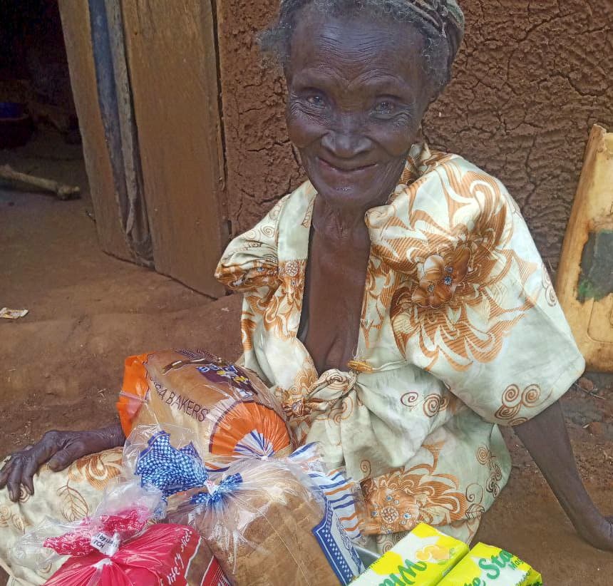 Elderly woman Marita sits smiling beside bags of delivered groceries including bread and produce.
