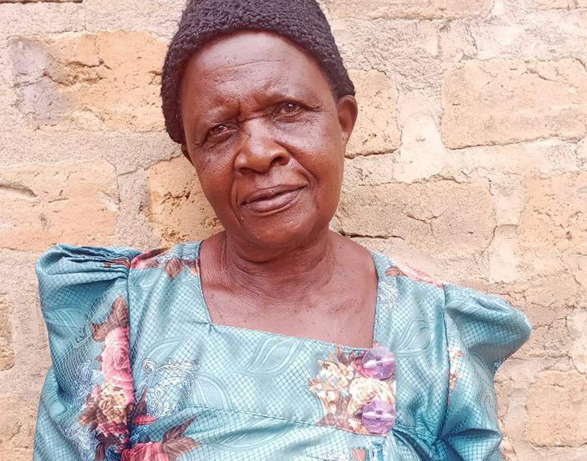 Portrait of an elderly woman, believed to be Nassozi Magret, wearing a headscarf and floral blouse against a brick wall.