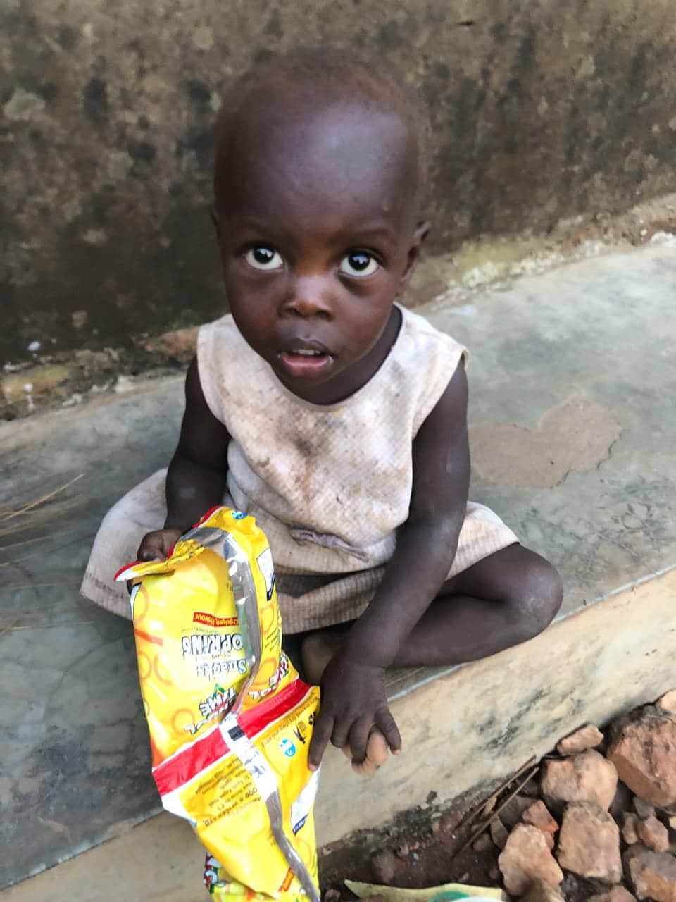 A young child sits on the ground holding a snack packet during a Miracle Medical Center outreach visit.