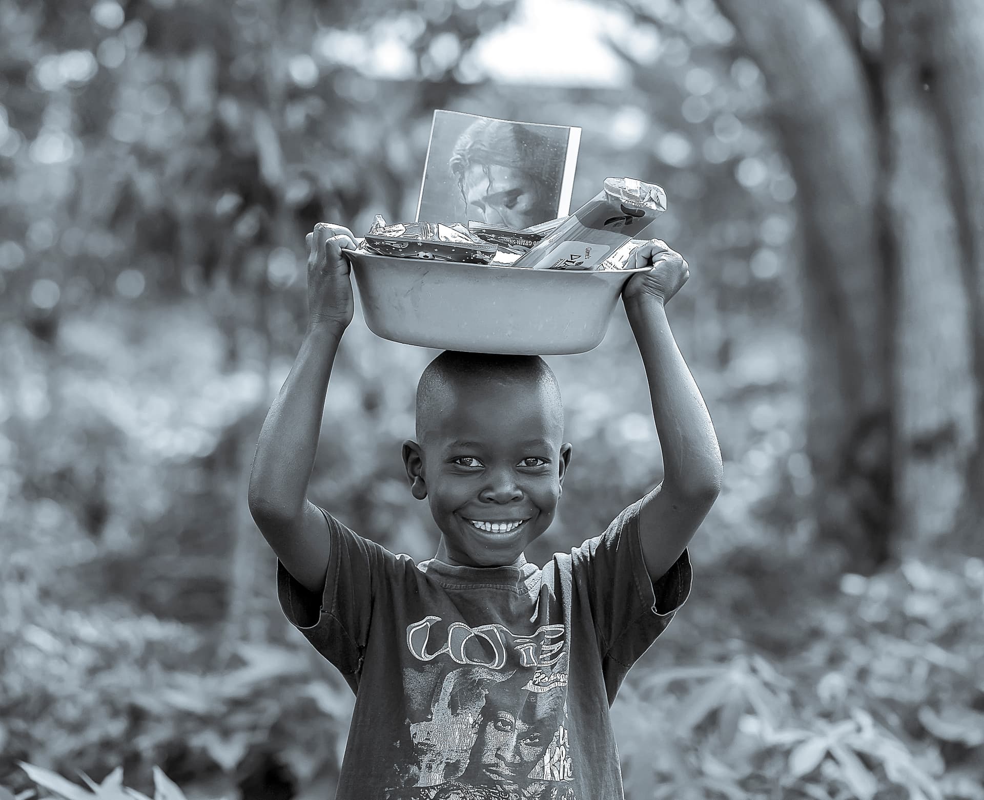 Black-and-white photo of a boy carrying a metal food basin balanced on his head.